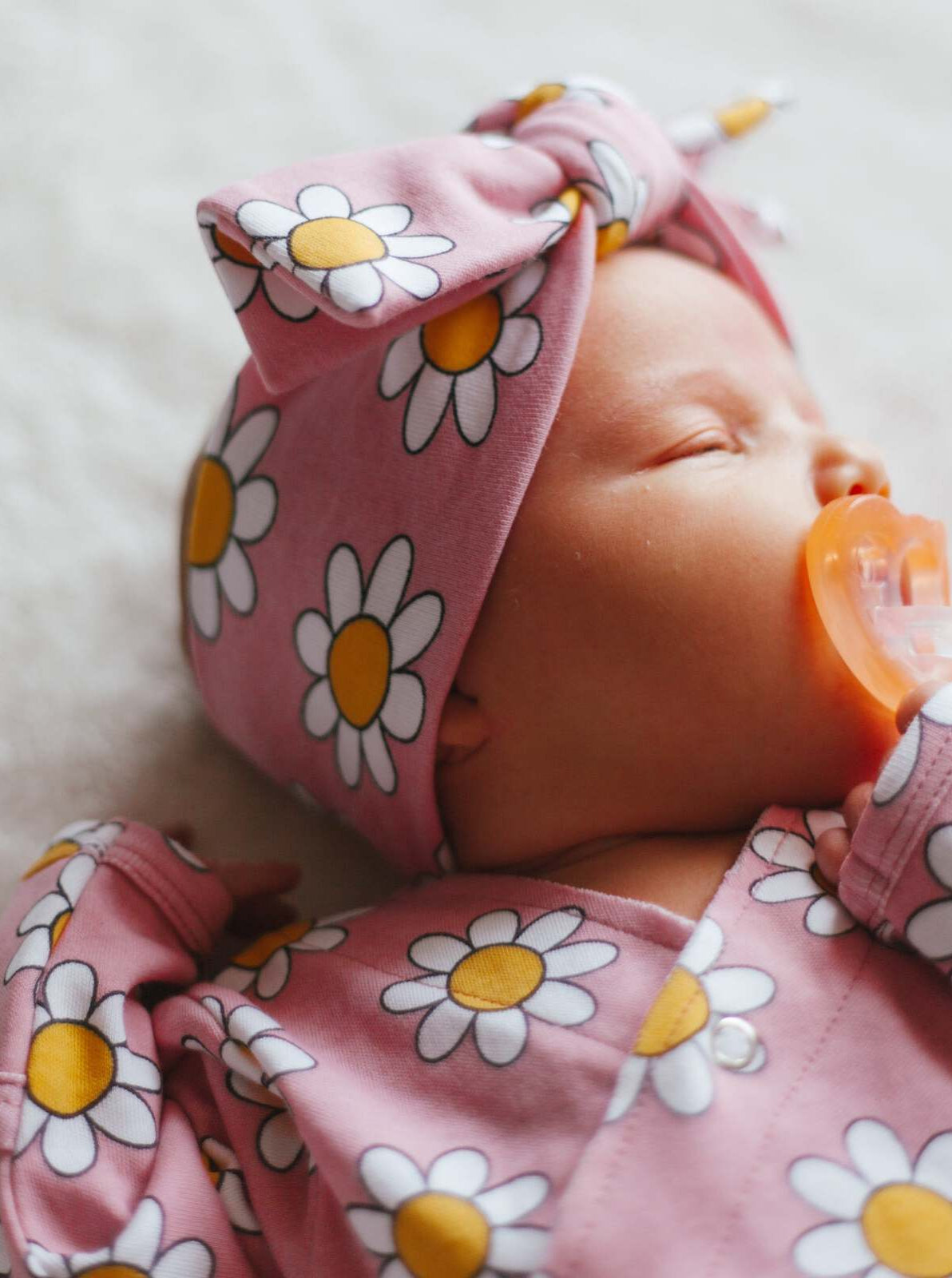 Infant sleeping peacefully, wearing a pink daisy-patterned outfit and a matching headband, with a pacifier.