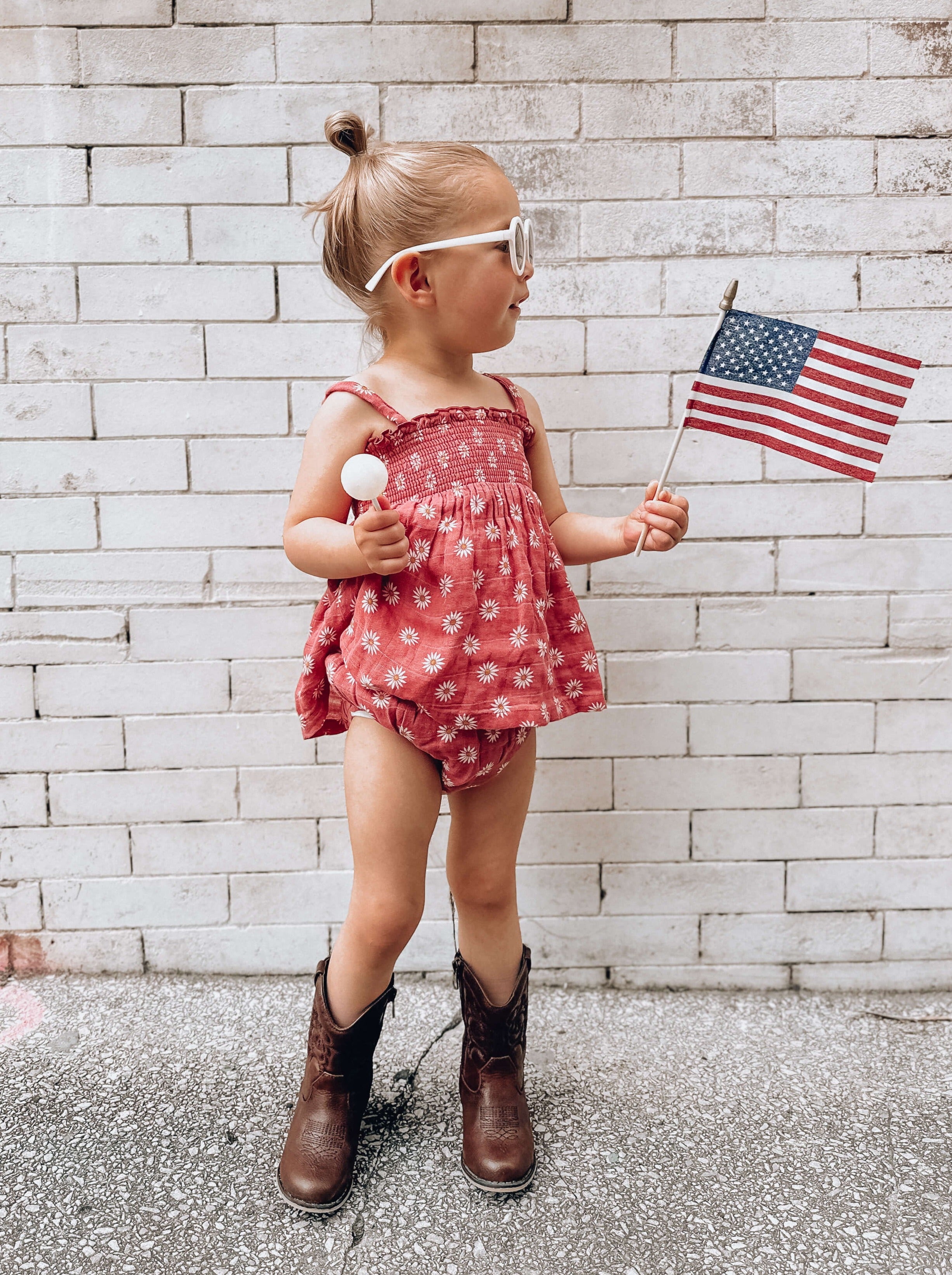 Girl in red floral outfit and brown boots holding a small American flag, smiling against a brick wall.