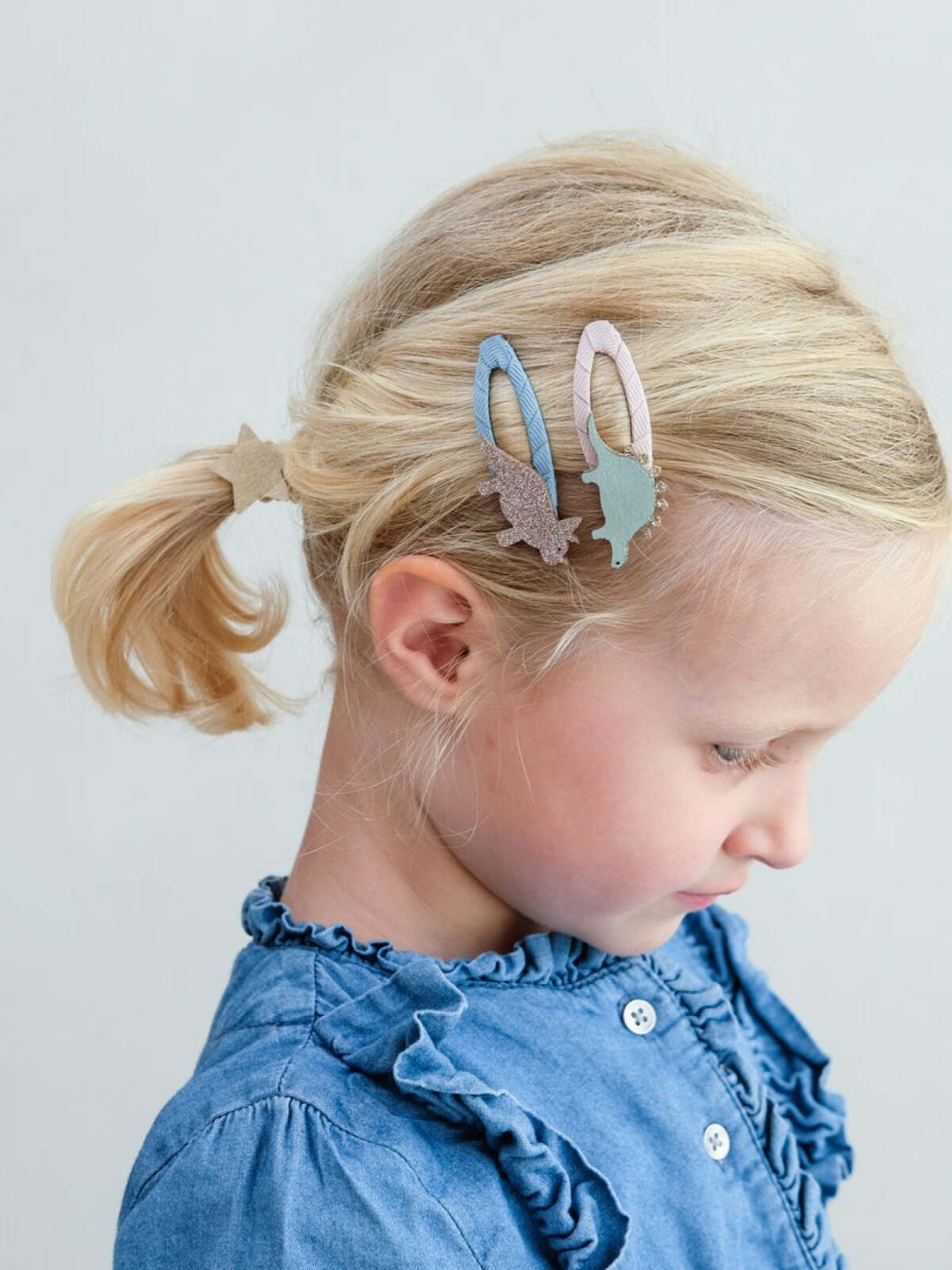 Girl with blonde hair, wearing a blue shirt and decorative hair clips, looking down against a light background.