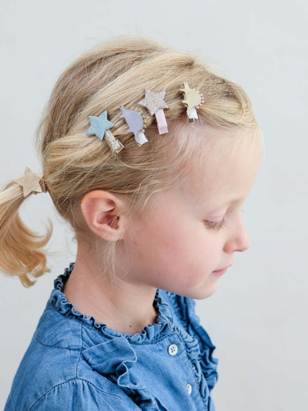 Young girl with blonde hair wearing star-shaped hair clips, side profile, in a light background.