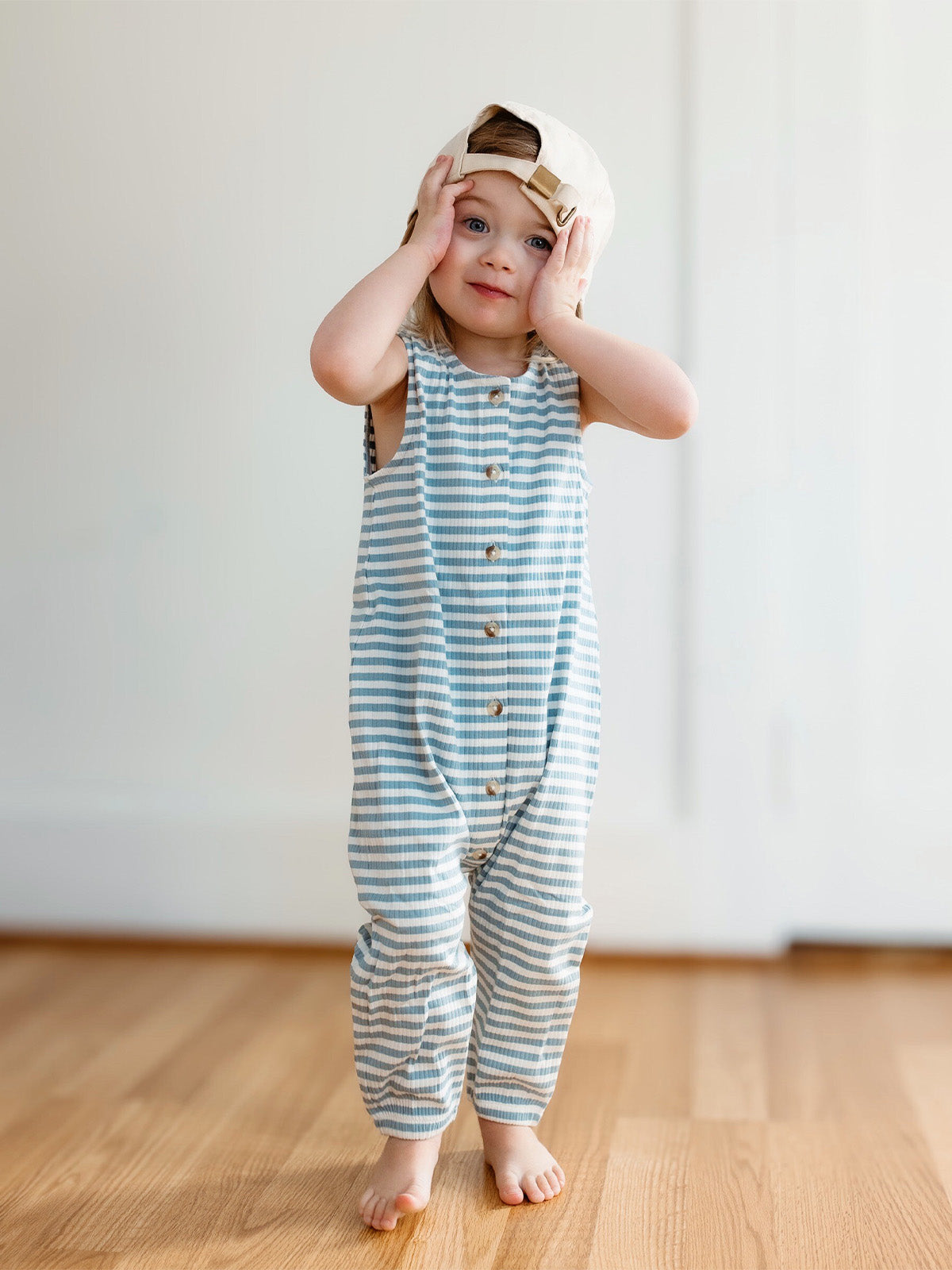 Young child in striped outfit and cap, smiling with hands on face, standing on wooden floor against a light background.