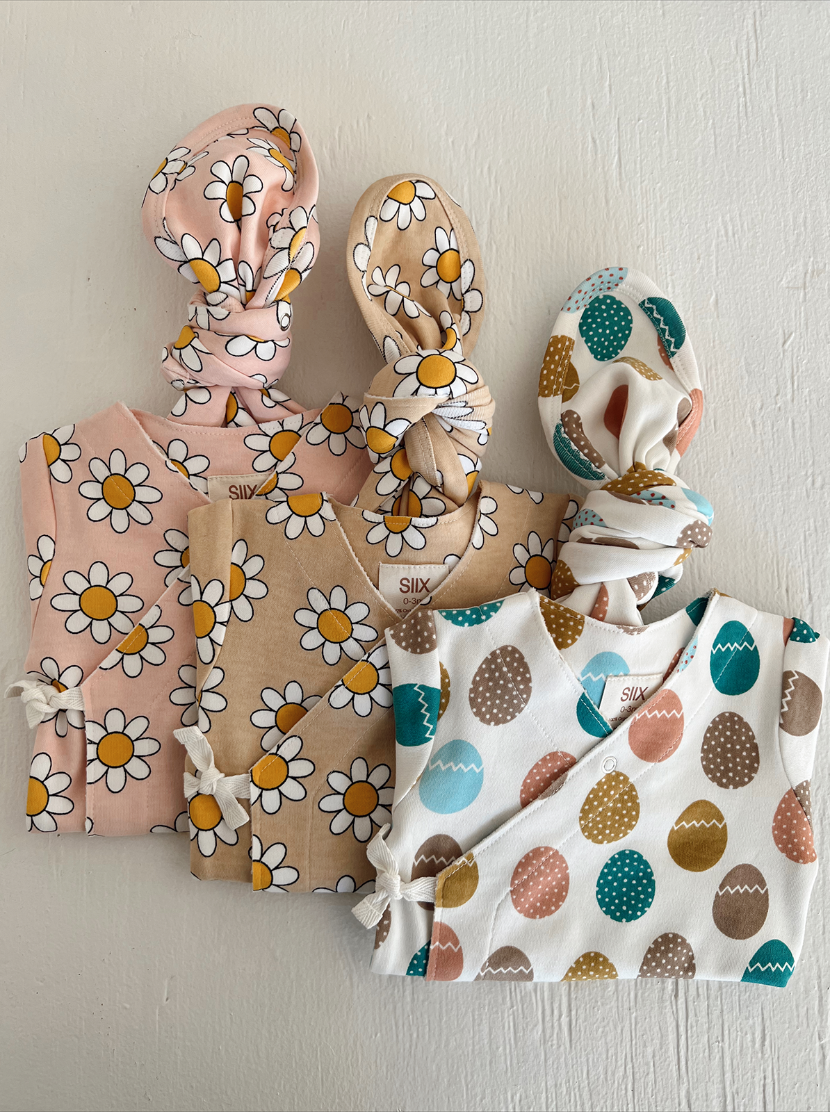 Three baby outfits with floral and egg patterns on a neutral background. Ideal for spring fashion.