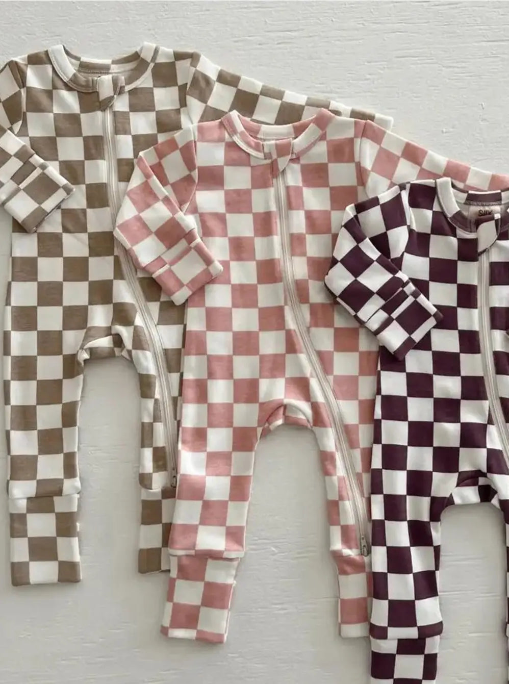 Three checkered baby rompers in brown, pink, and burgundy on a light wooden surface.