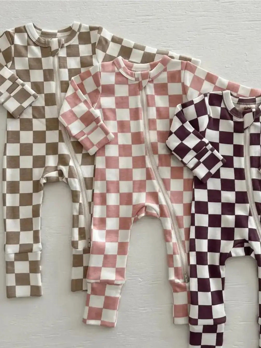 Three checkered baby rompers in brown, pink, and burgundy on a light wooden surface.