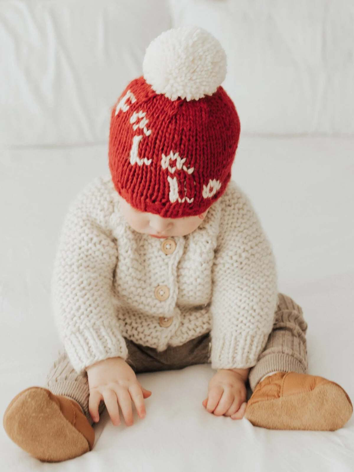 Baby wearing a red knitted hat with a pom-pom and a cream cardigan, sitting on a bed, playing with hands.