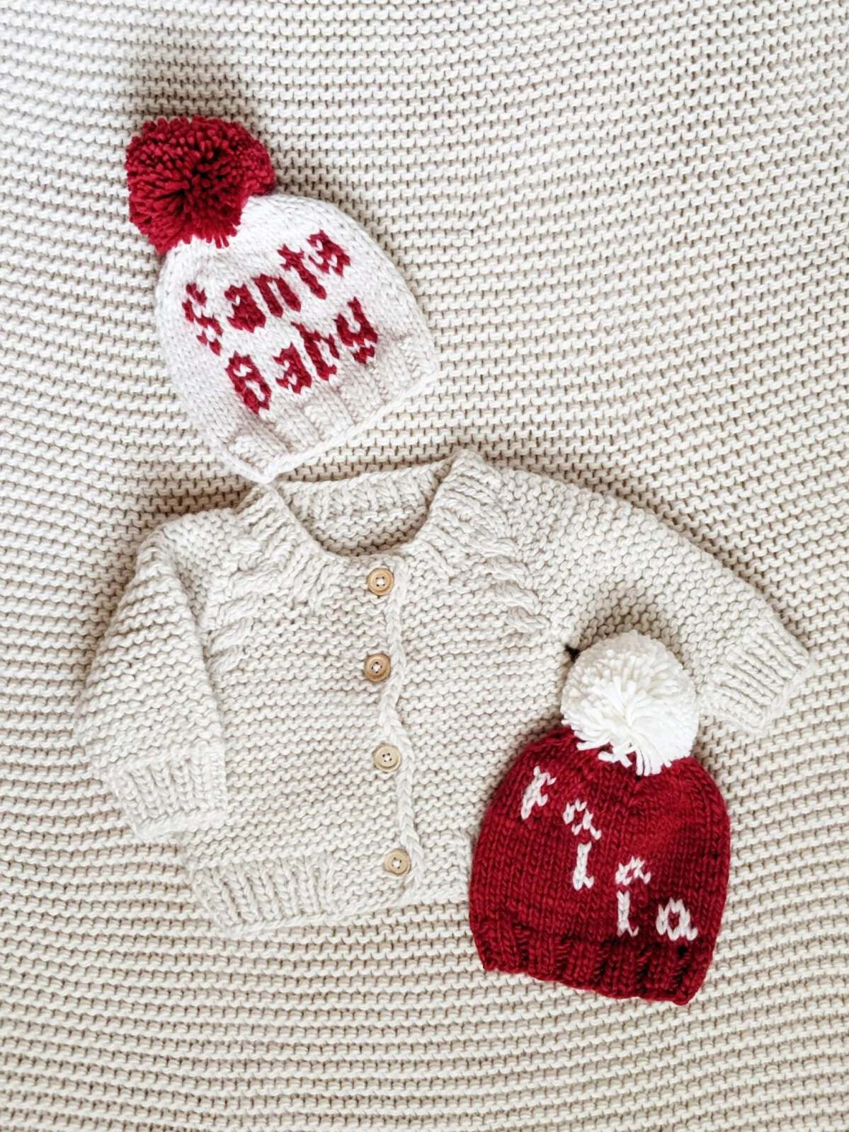 Knit toddler sweater and two beanies on a textured blanket, one saying "Santa Baby" and the other "tia."