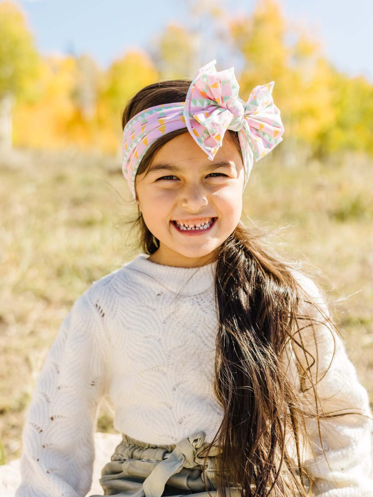 Smiling girl with long hair wearing a colorful headband, seated outdoors with autumn trees in the background.