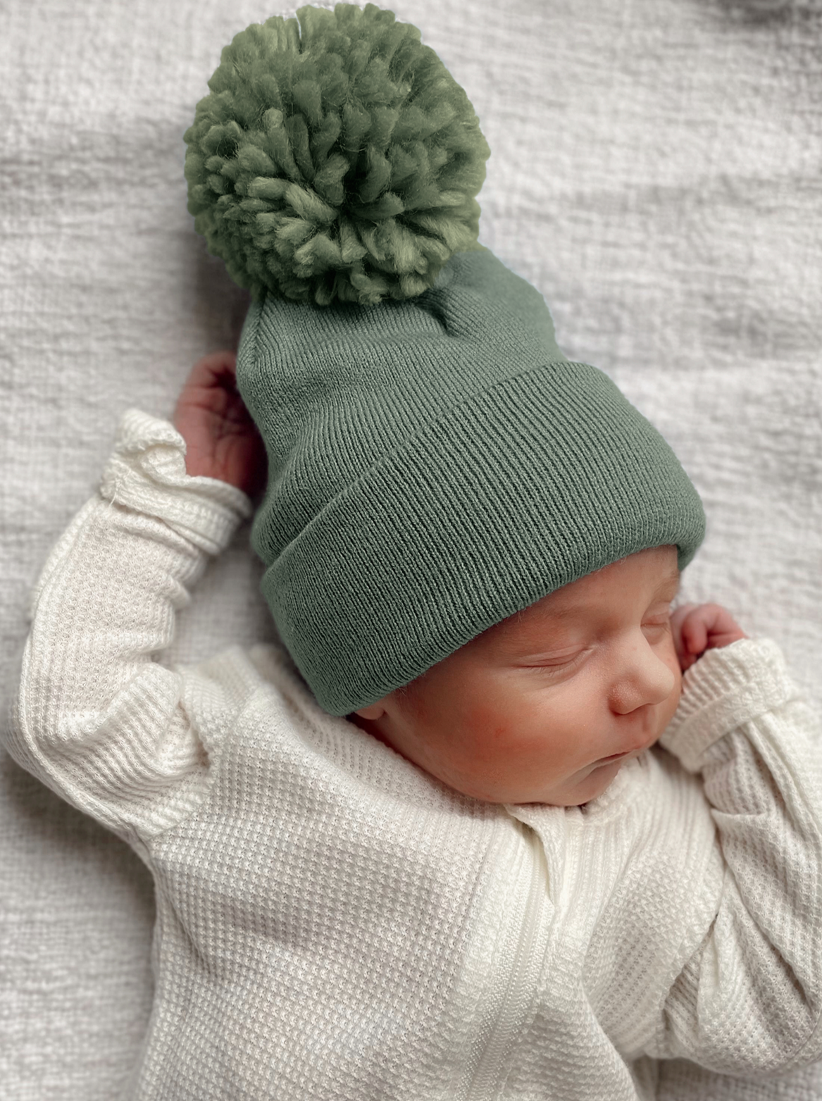 Newborn asleep in a green pom-pom hat, wearing a white cozy outfit, resting on a textured gray blanket.