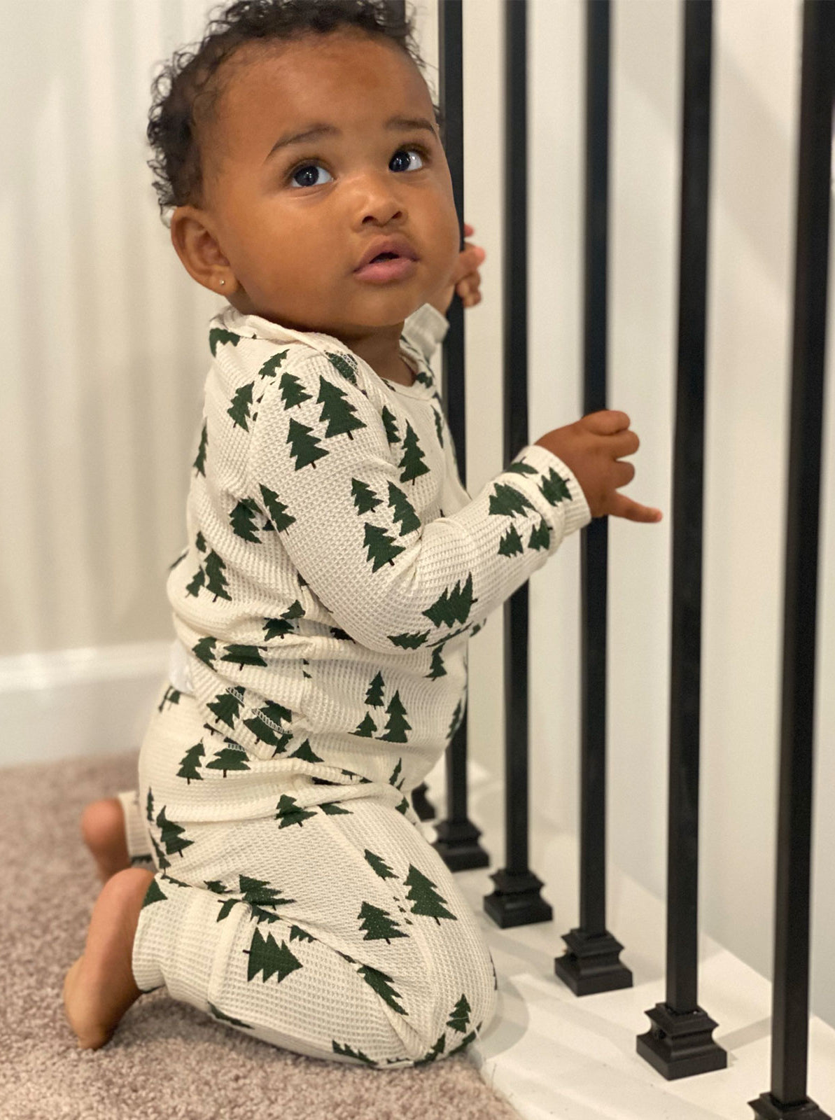 Infant in festive pajamas, kneeling by black railing, looking curiously at the camera.