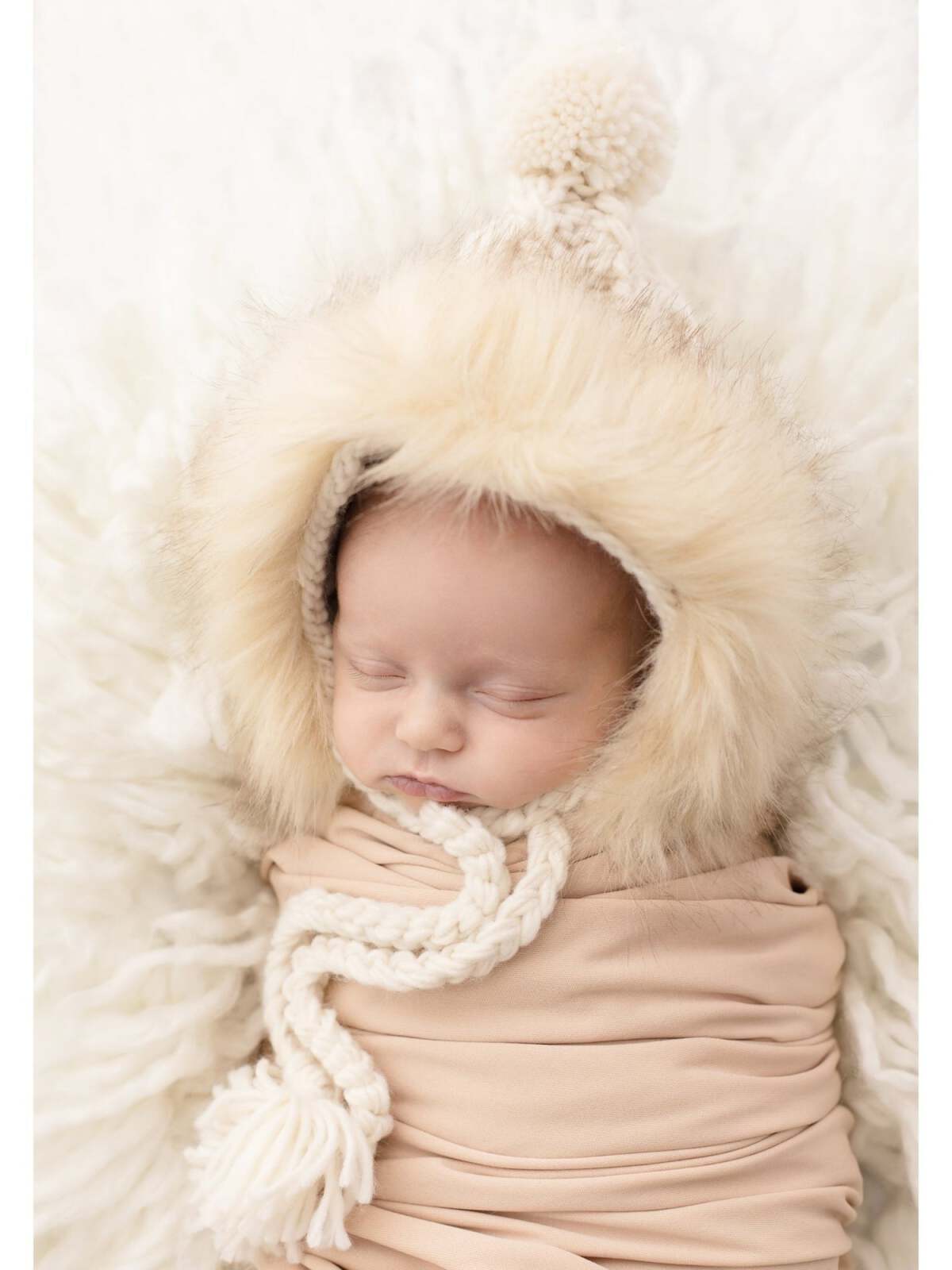 Sleeping baby wrapped in beige swaddle, wearing a cozy fur-lined hood with a pom-pom, on a fluffy white blanket.