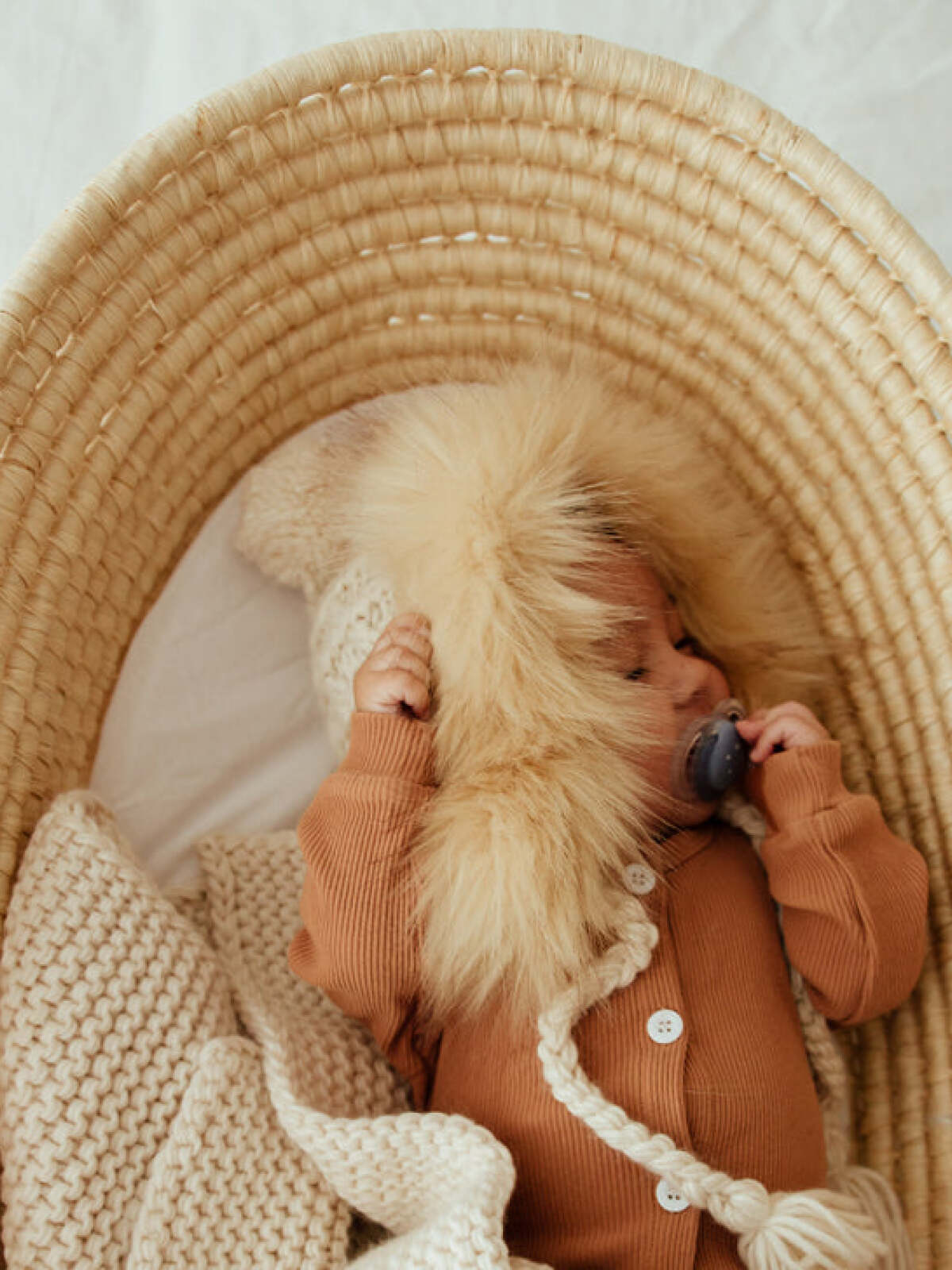 Baby in a cozy woven basket, wearing a furry hood and holding a pacifier, surrounded by soft blankets.