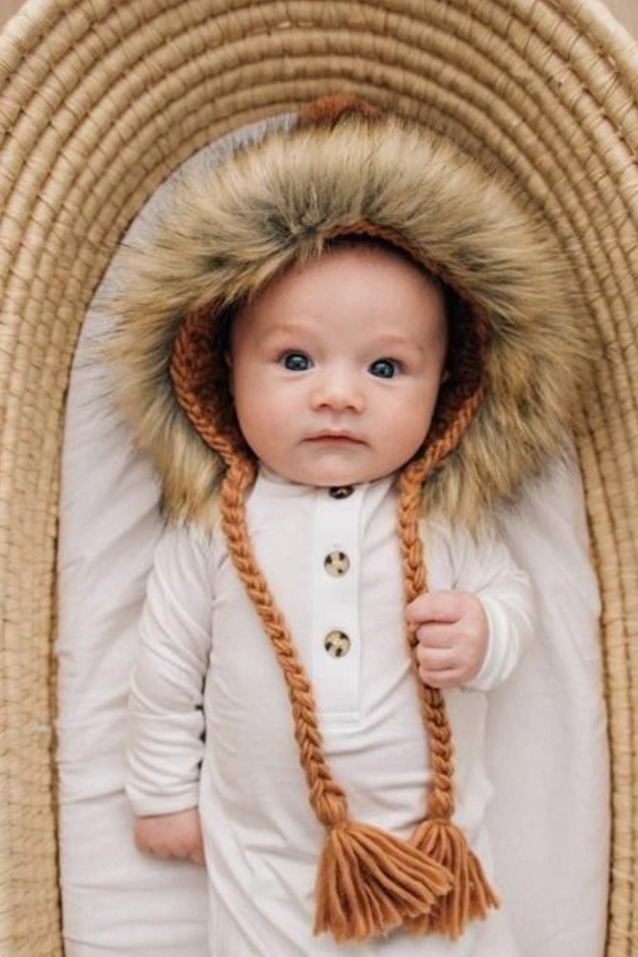 Baby wearing a fluffy hooded outfit, lying in a woven basket, with a curious expression.