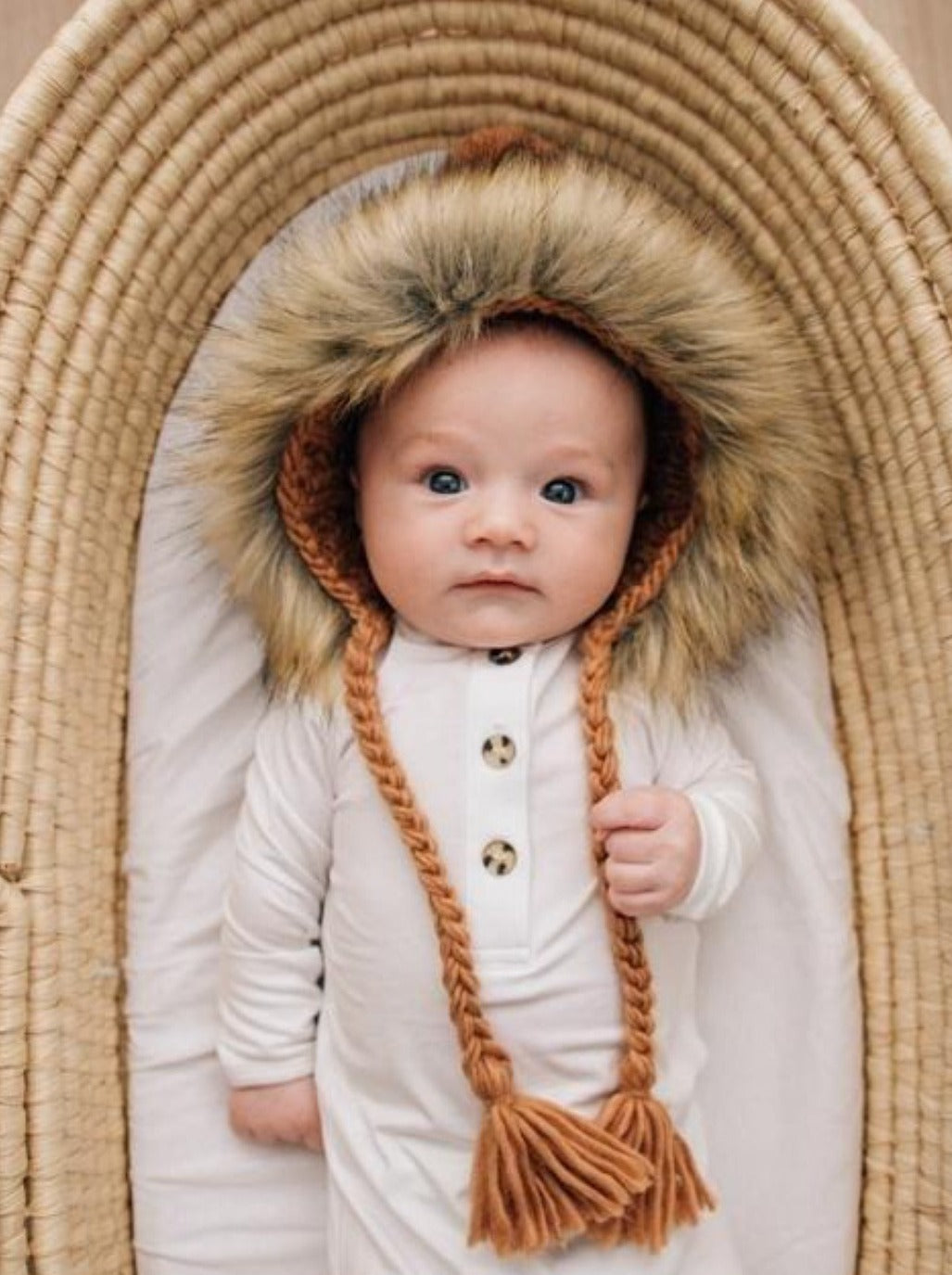 Baby wearing a fluffy hooded outfit, lying in a woven basket, with a curious expression.