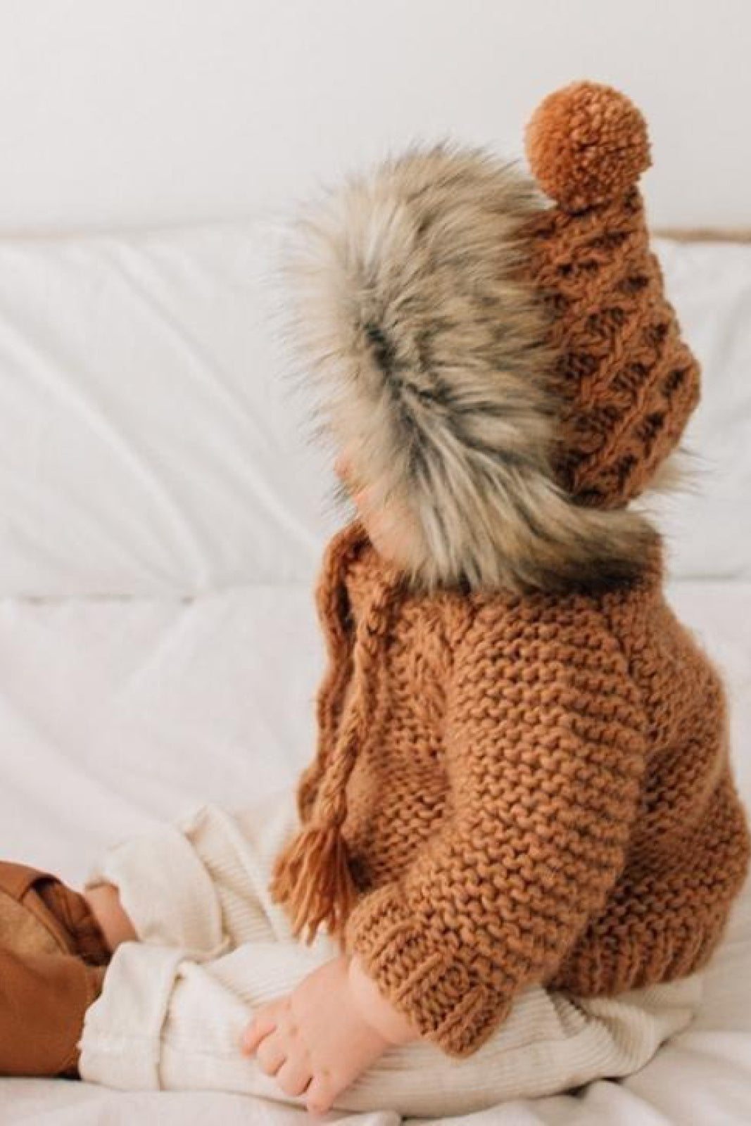 Toddler in a cozy brown knitted sweater and hat with a fur trim, sitting on a bed.