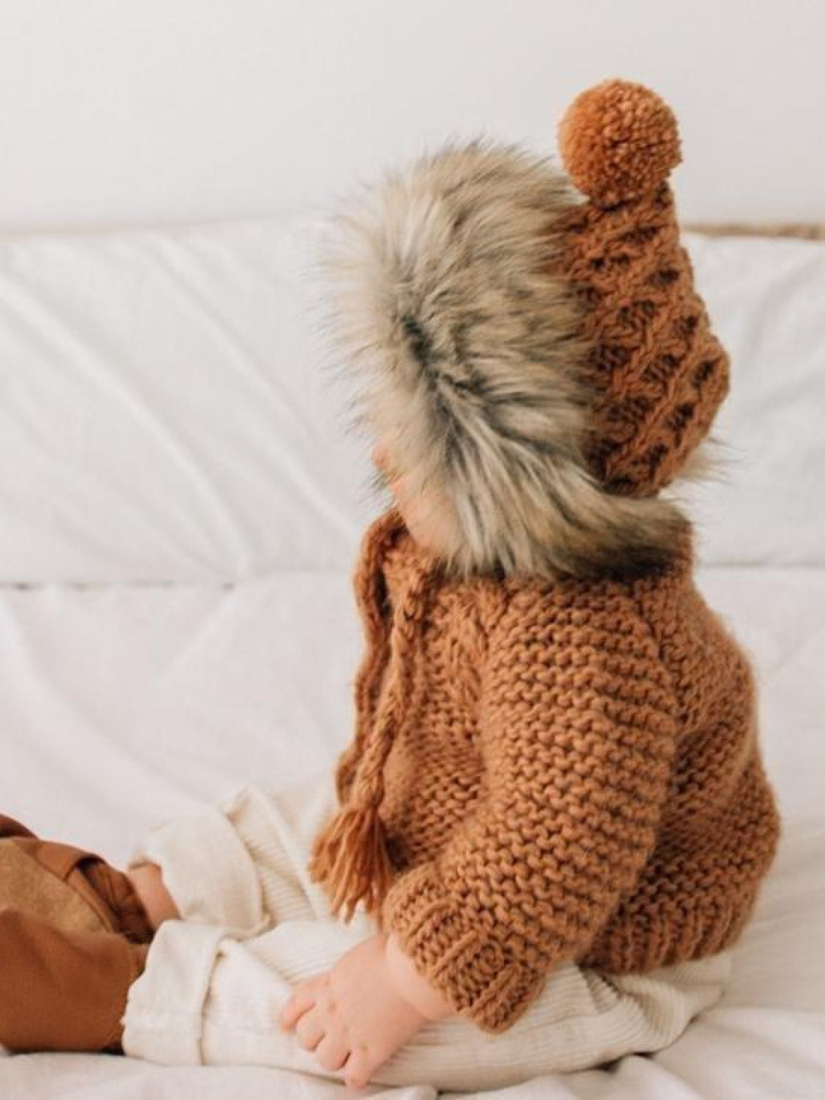 Toddler in a cozy brown knitted sweater and hat with a fur trim, sitting on a bed.