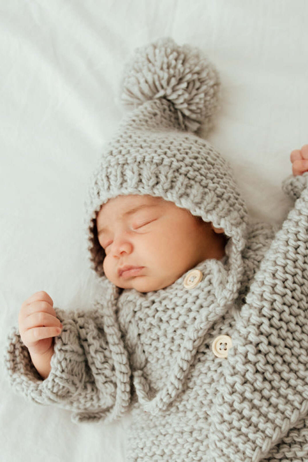 Baby sleeping peacefully in a cozy, knitted gray outfit with a pom-pom hat, resting on a soft surface.