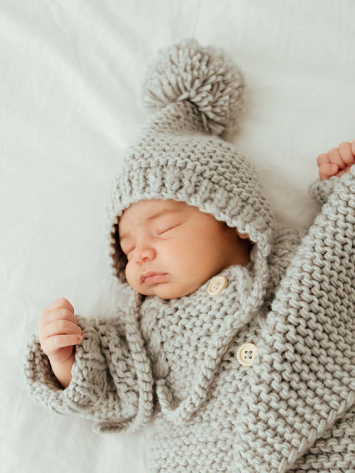 Baby sleeping peacefully in a cozy, knitted gray outfit with a pom-pom hat, resting on a soft surface.