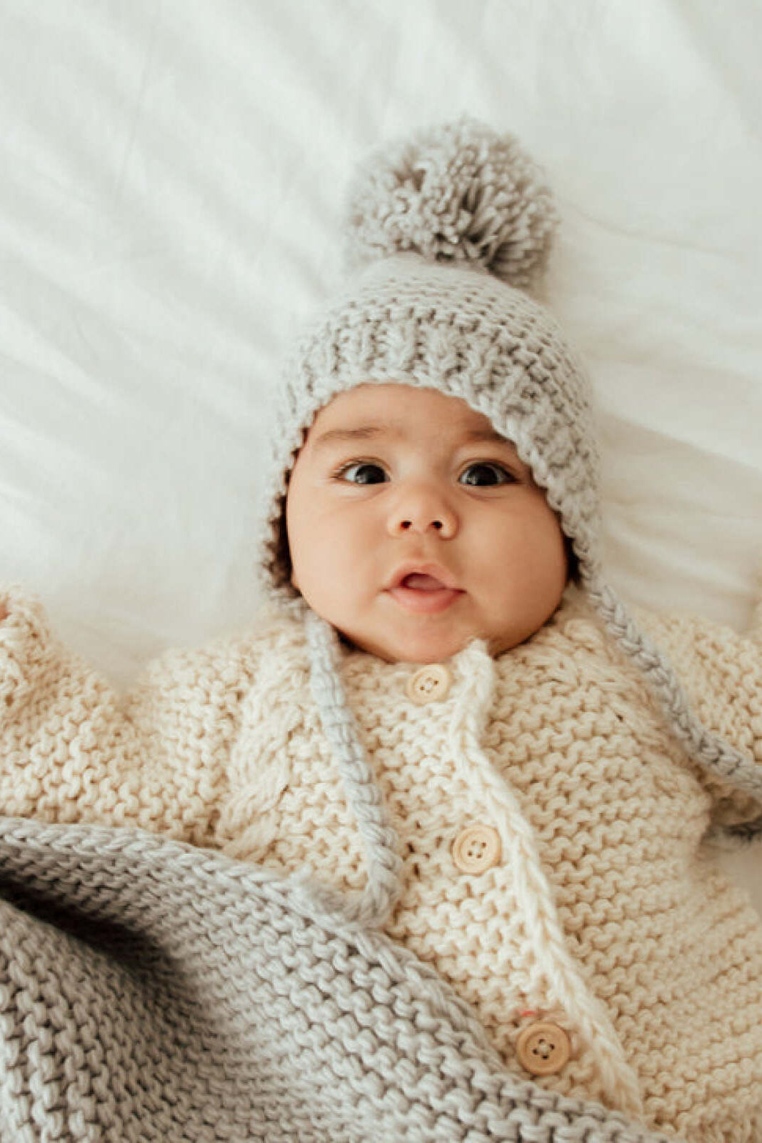 Baby wearing a knitted hat and cozy sweater, lying on a white blanket, looking up with a playful expression.