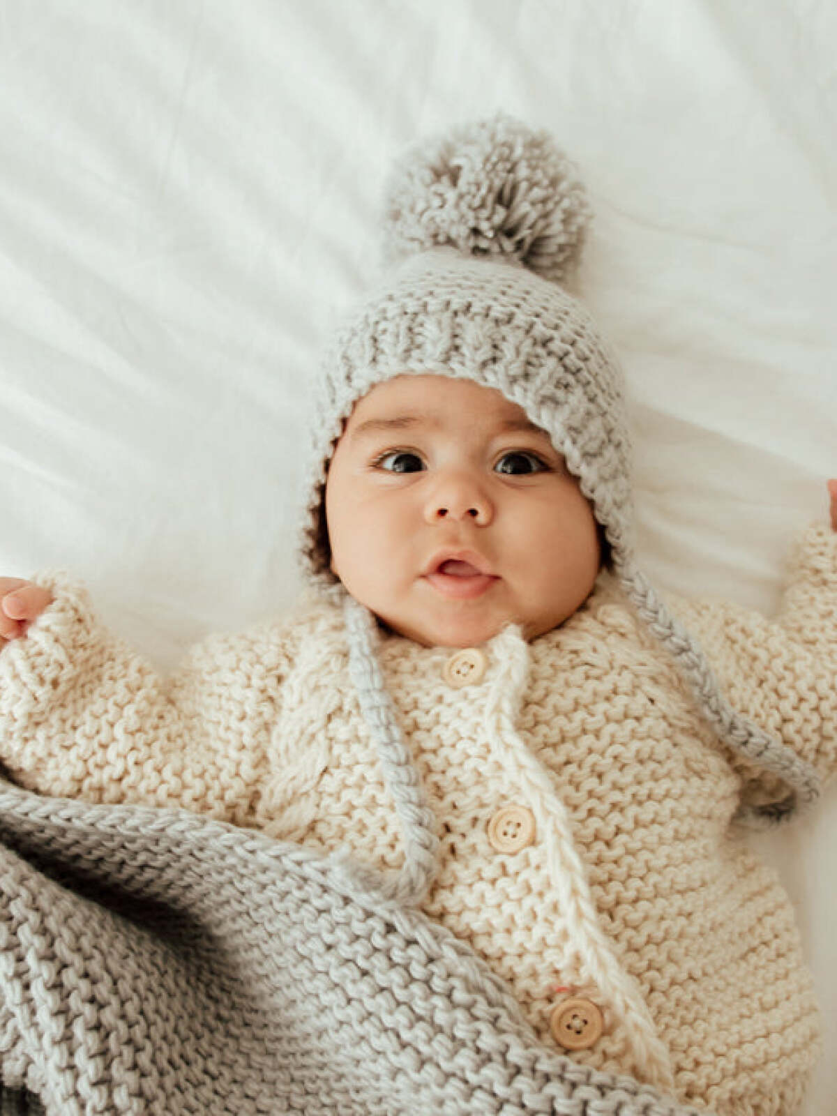 Baby wearing a knitted hat and cozy sweater, lying on a white blanket, looking up with a playful expression.