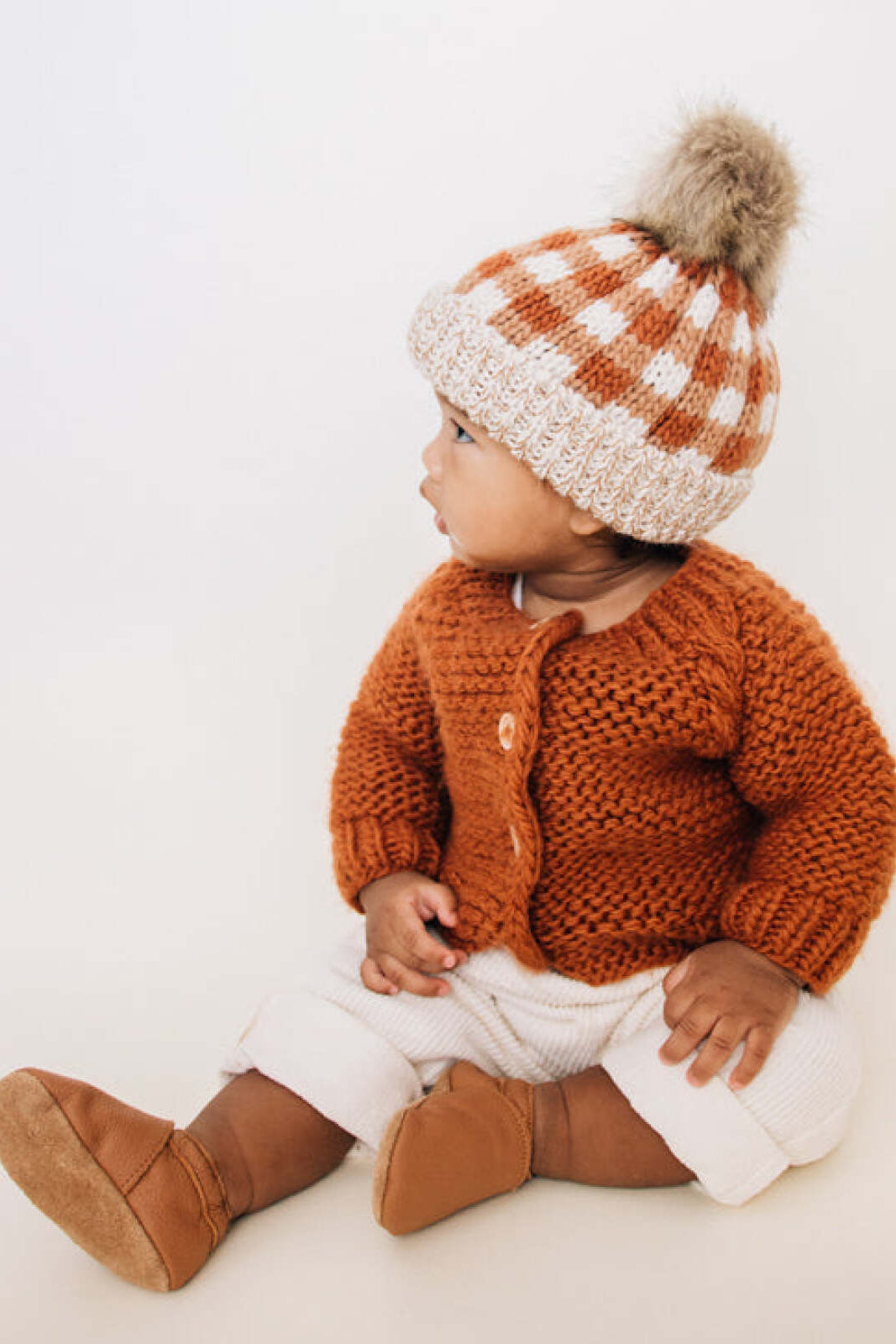 Infant wearing a knitted orange cardigan and checkered hat, sitting on a light background.