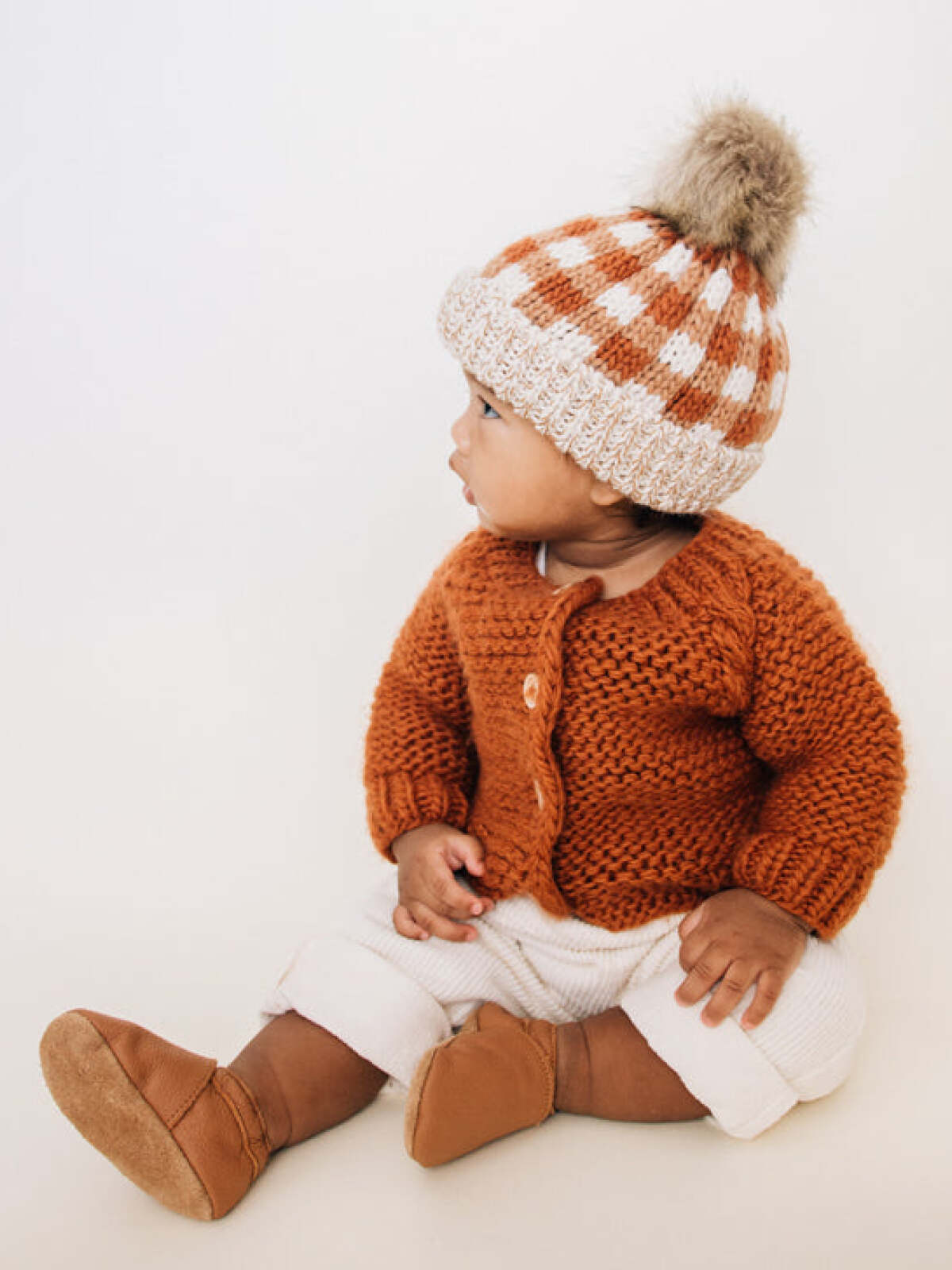 Infant wearing a knitted orange cardigan and checkered hat, sitting on a light background.