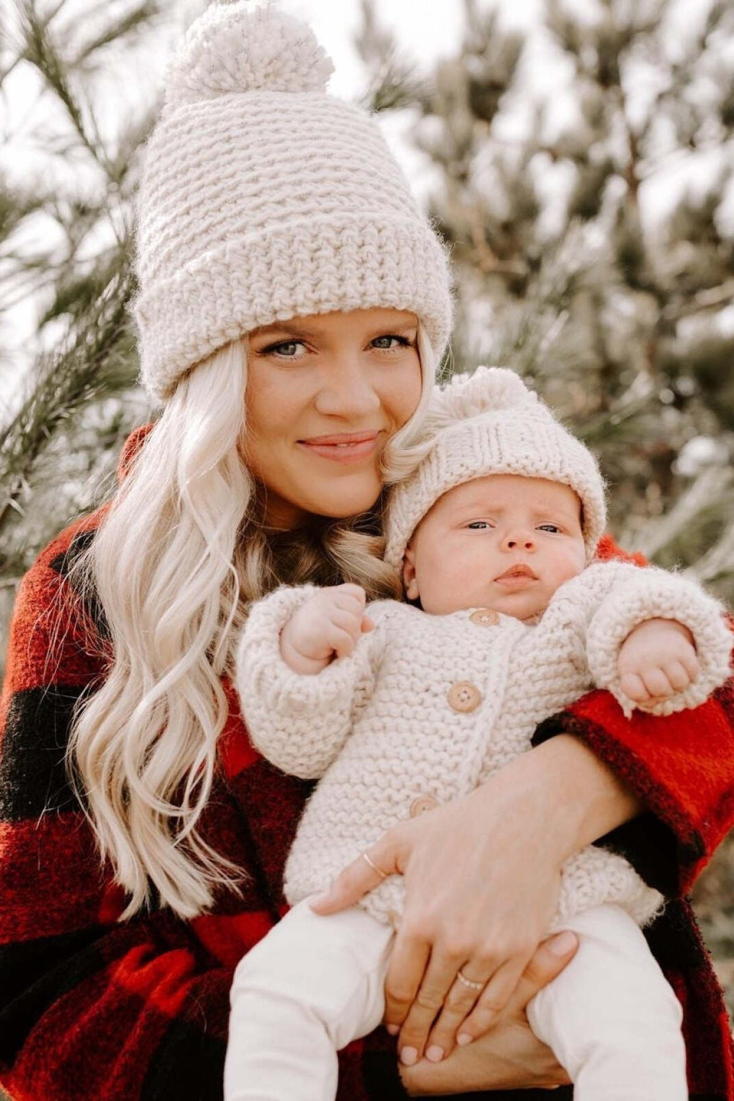 Woman holds an infant in cozy winter outfits, surrounded by pine trees. Soft lighting highlights their warmth and connection.