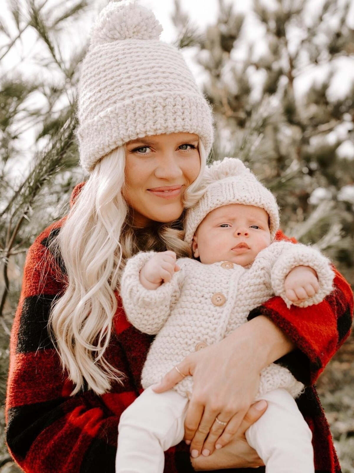 Woman holds an infant in cozy winter outfits, surrounded by pine trees. Soft lighting highlights their warmth and connection.