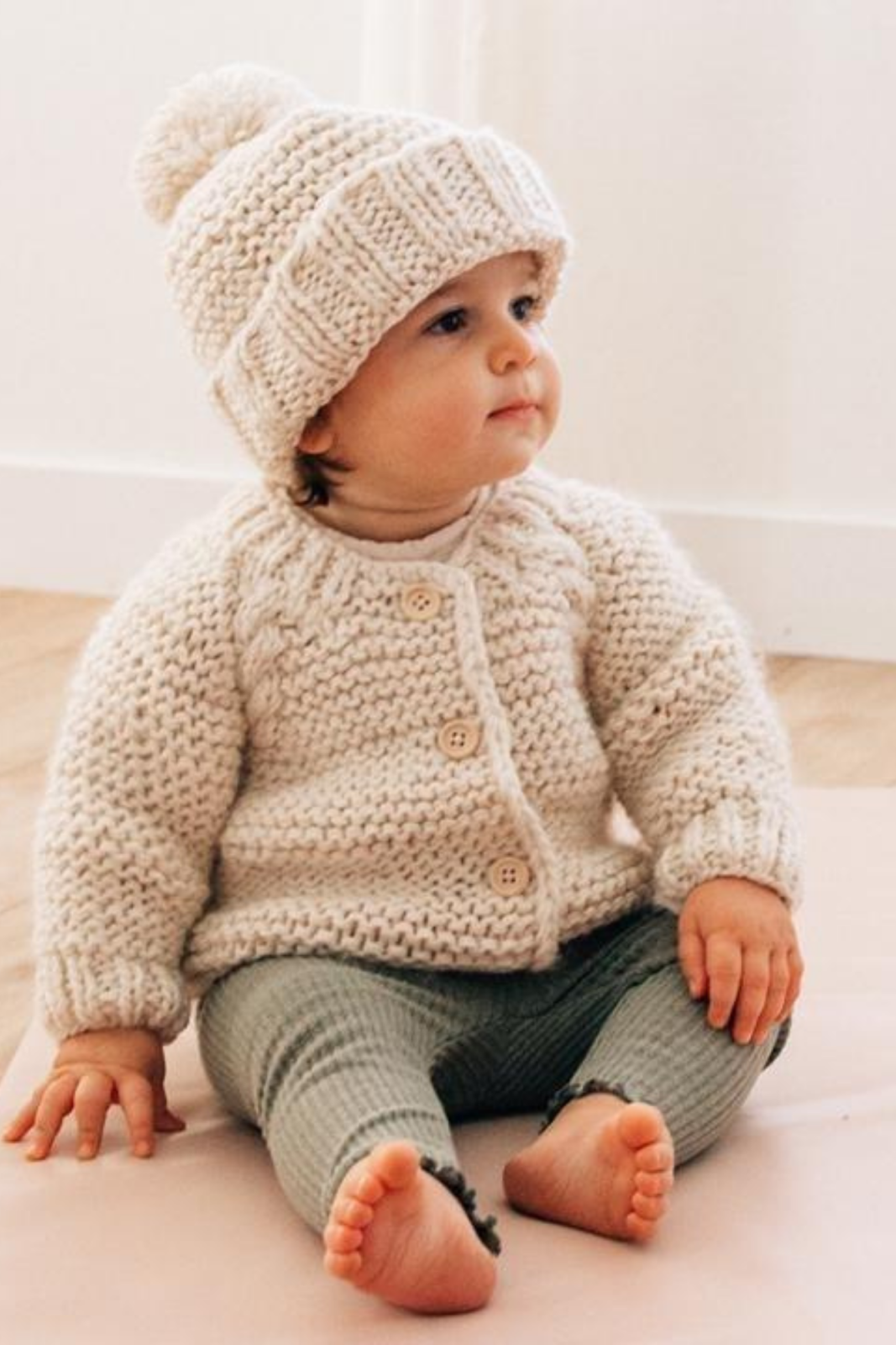 Toddler in cozy cream sweater and hat, sitting on soft surface with wooden floor background.