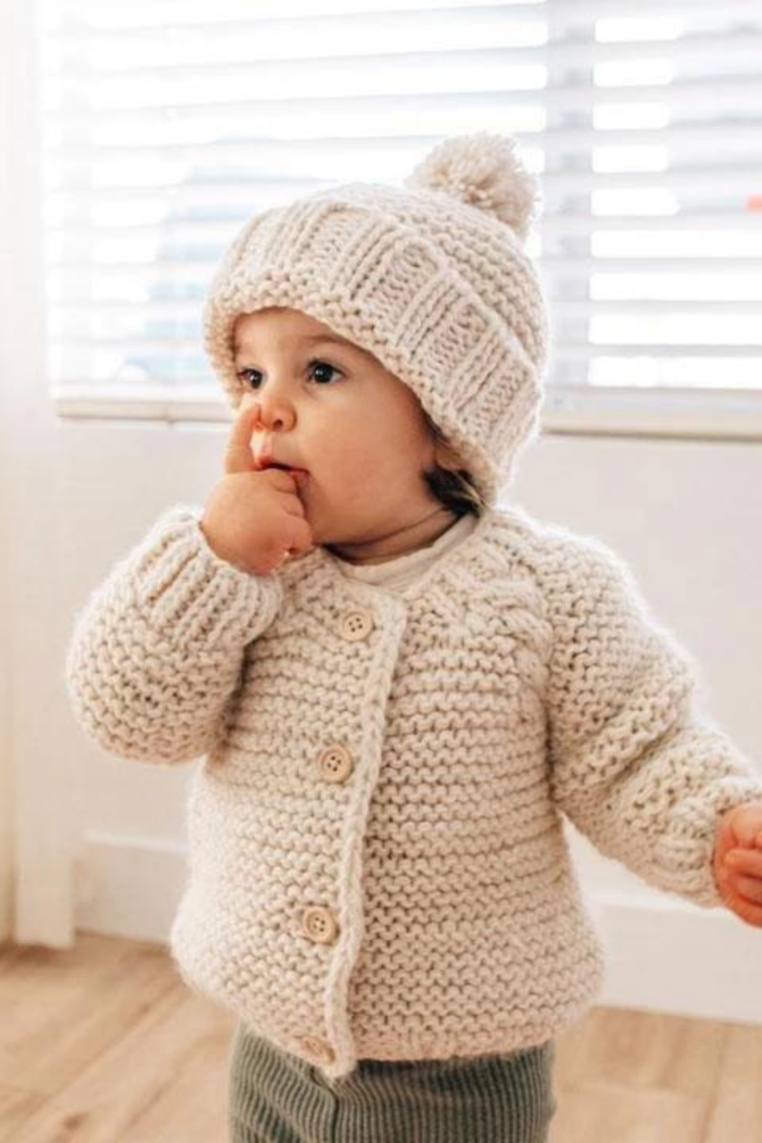 Toddler in a cozy knitted sweater and hat, curiously sucking their finger by a window with blinds.