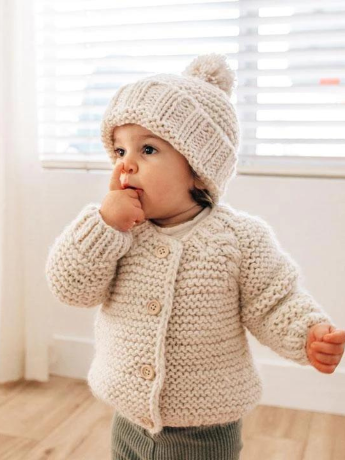 Toddler in a cozy knitted sweater and hat, curiously sucking their finger by a window with blinds.