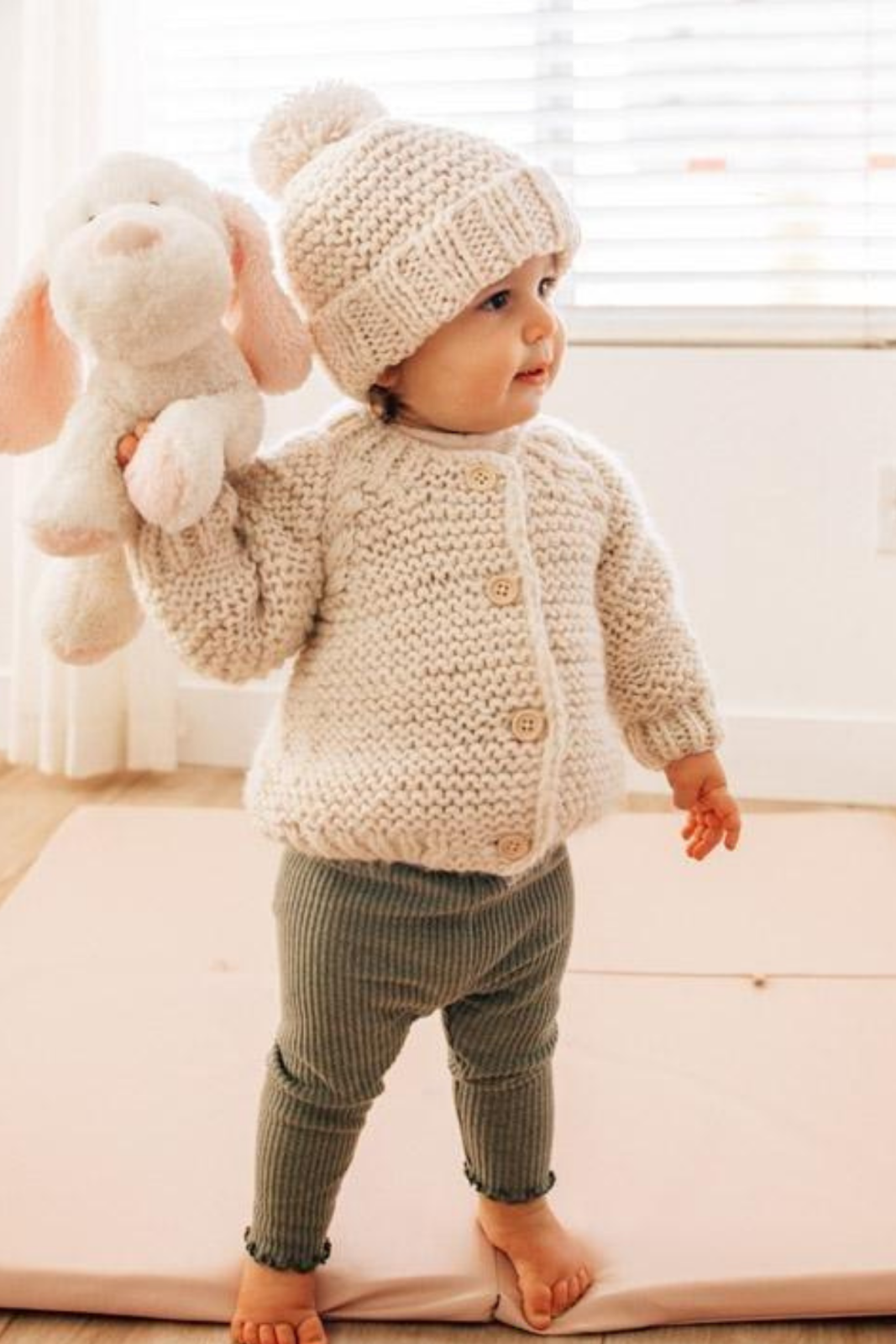 Toddler in a cozy knitted outfit holding a stuffed animal, standing on a soft mat in a bright indoor space.