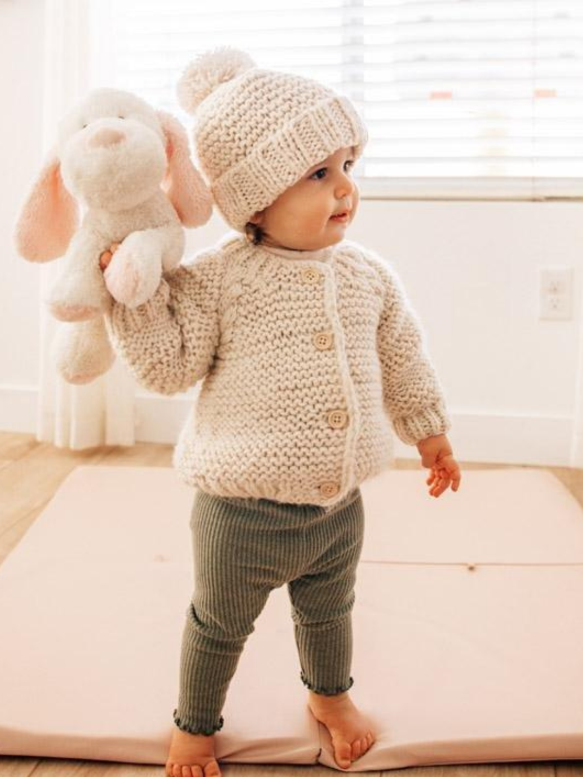 Toddler in a cozy knitted outfit holding a stuffed animal, standing on a soft mat in a bright indoor space.