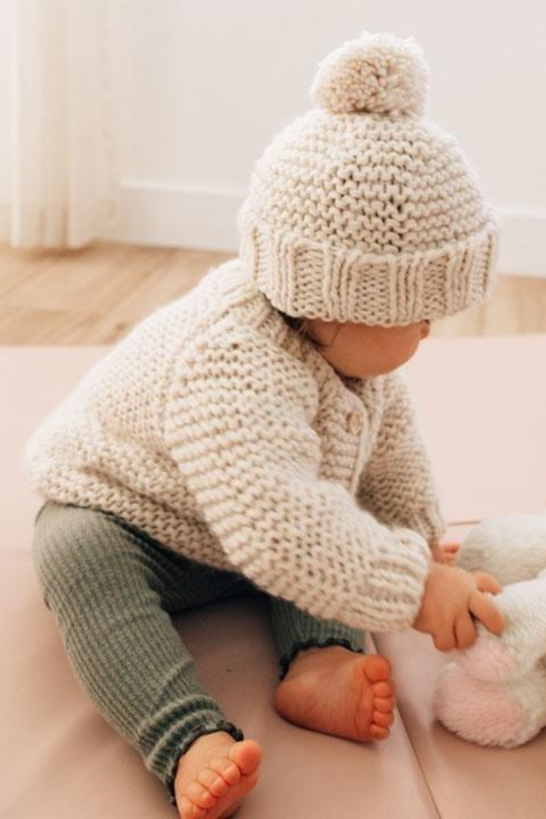 Baby in a knitted sweater and hat sits on the floor, playing with a plush toy. Soft, cozy atmosphere.