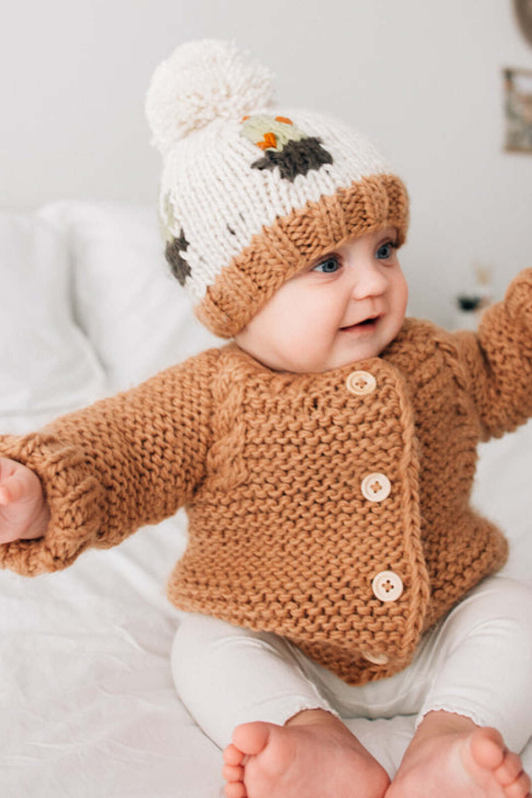 Baby in cozy knitted sweater and pom-pom hat, smiling on a soft bedspread.