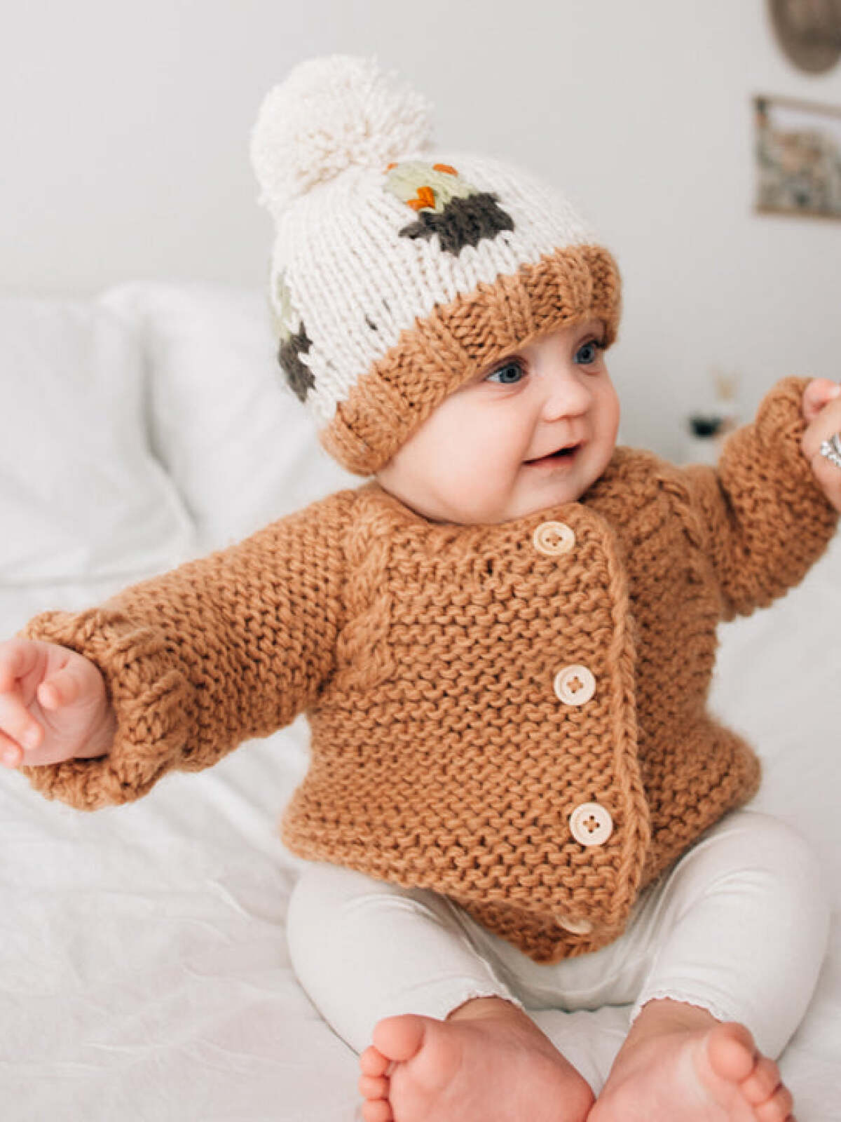 Baby in cozy knitted sweater and pom-pom hat, smiling on a soft bedspread.
