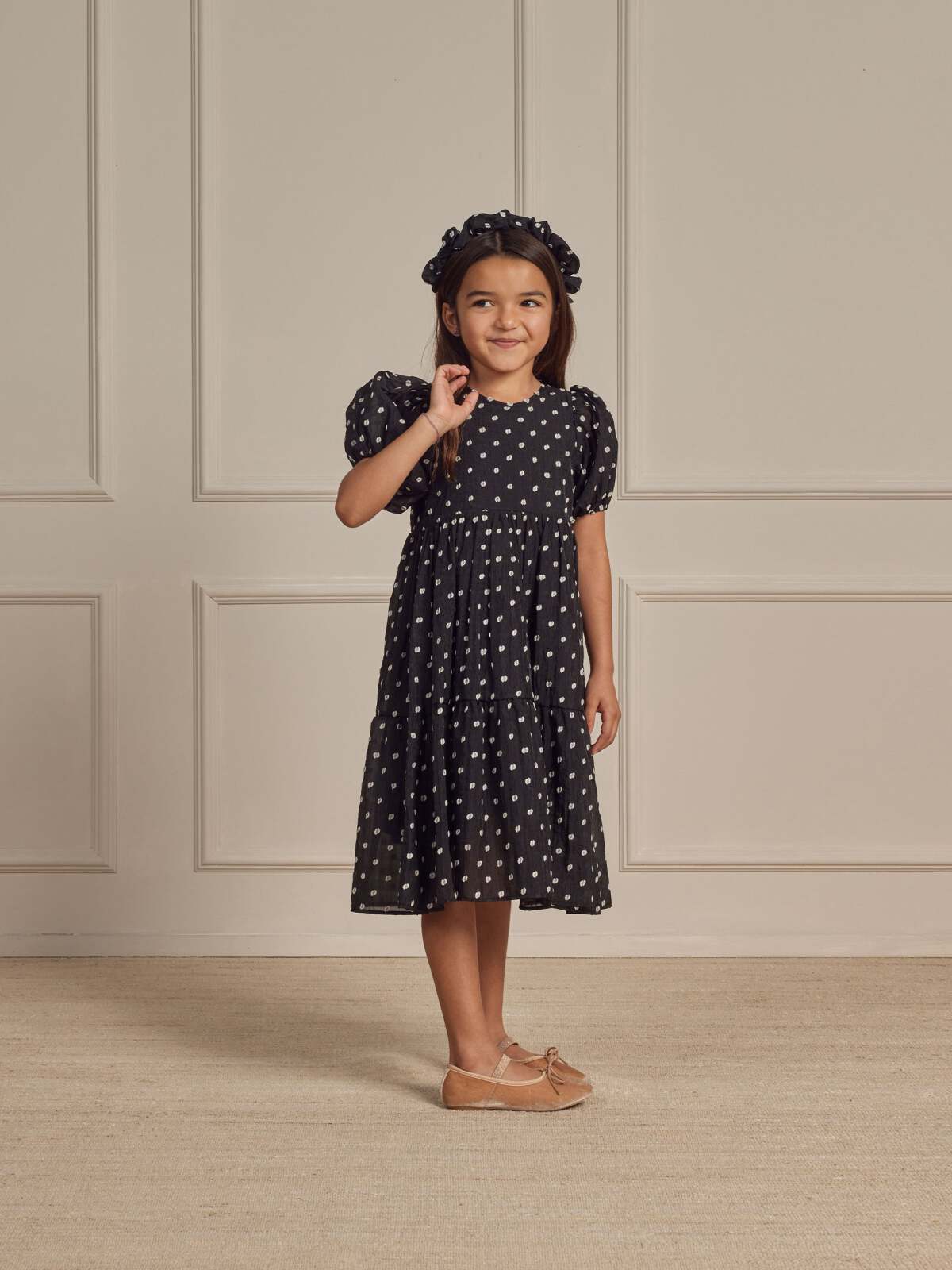 Girl in a polka dot dress and headband smiles while standing against a neutral wall backdrop.