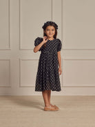 Girl in a polka dot dress and headband smiles while standing against a neutral wall backdrop.