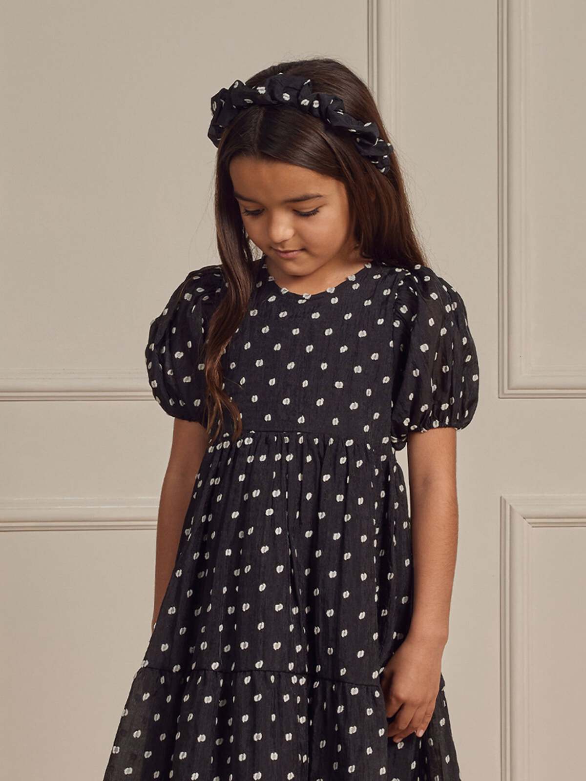 Girl in a black polka dot dress with puff sleeves and matching headband, standing in a neutral-toned room.