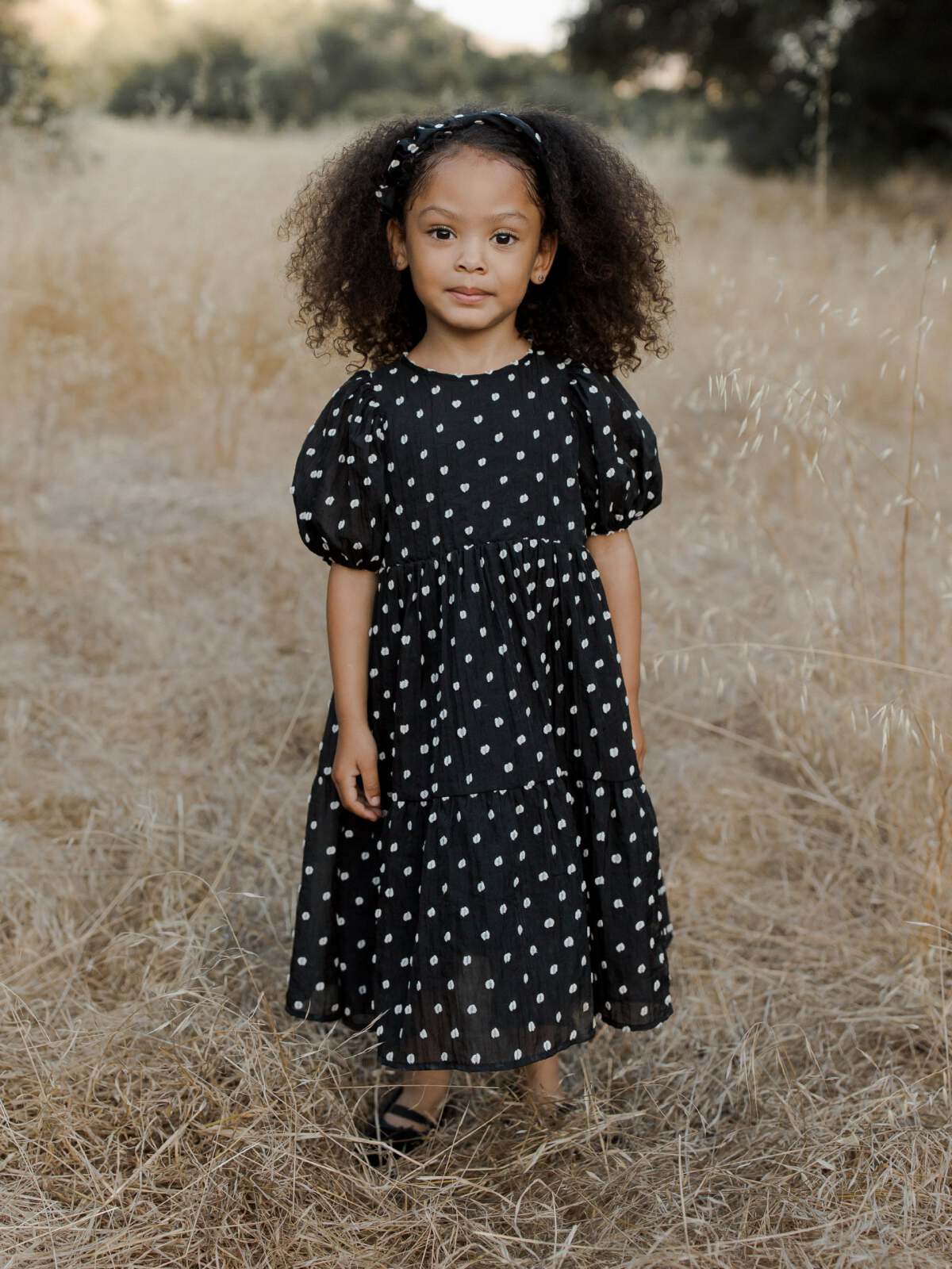 Child in a black polka dot dress standing in a field of dry grass, smiling at the camera.