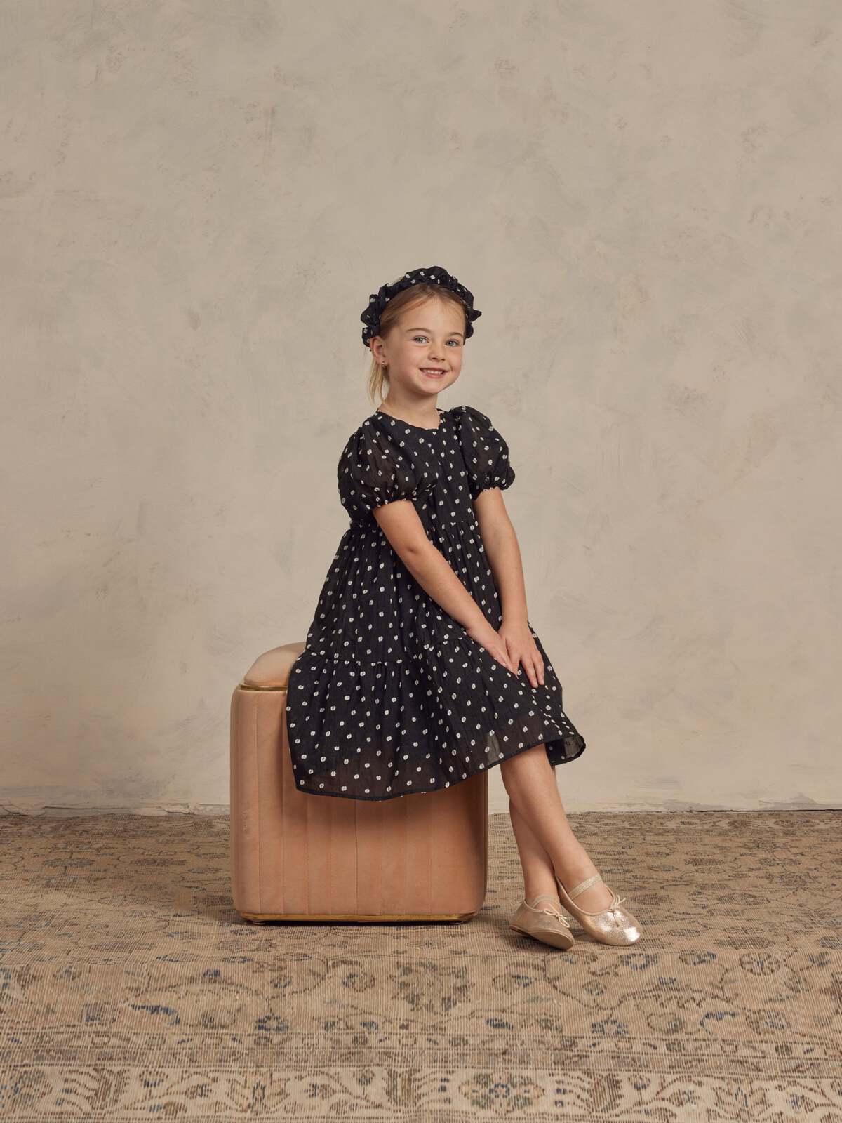 Girl in a polka dot dress and ballet shoes sitting on a beige stool against a textured background.