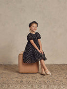 Girl in a polka dot dress and ballet shoes sitting on a beige stool against a textured background.