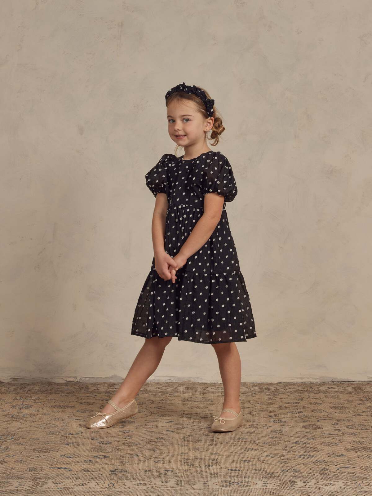 Girl in a polka dot dress and headband stands smiling on a textured background, showcasing a playful fashion look.