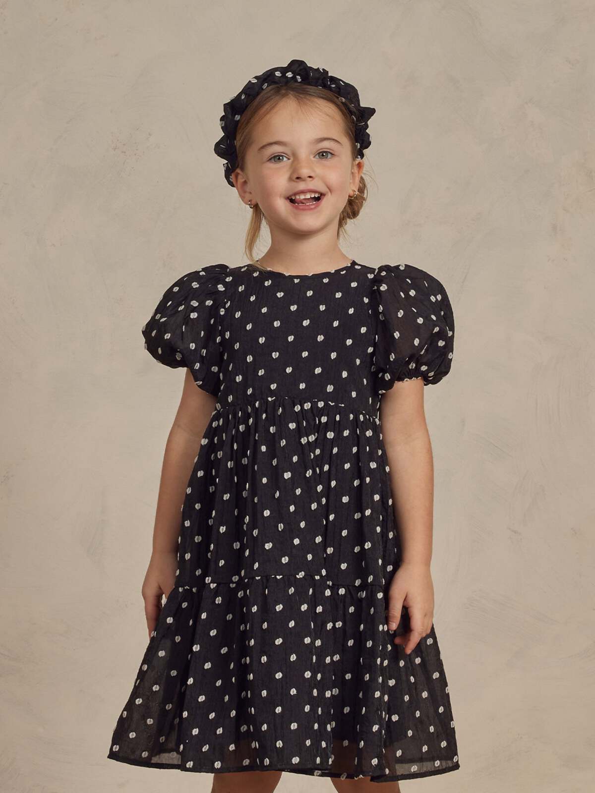 Smiling girl wearing a black polka dot dress and matching headband against a neutral background.