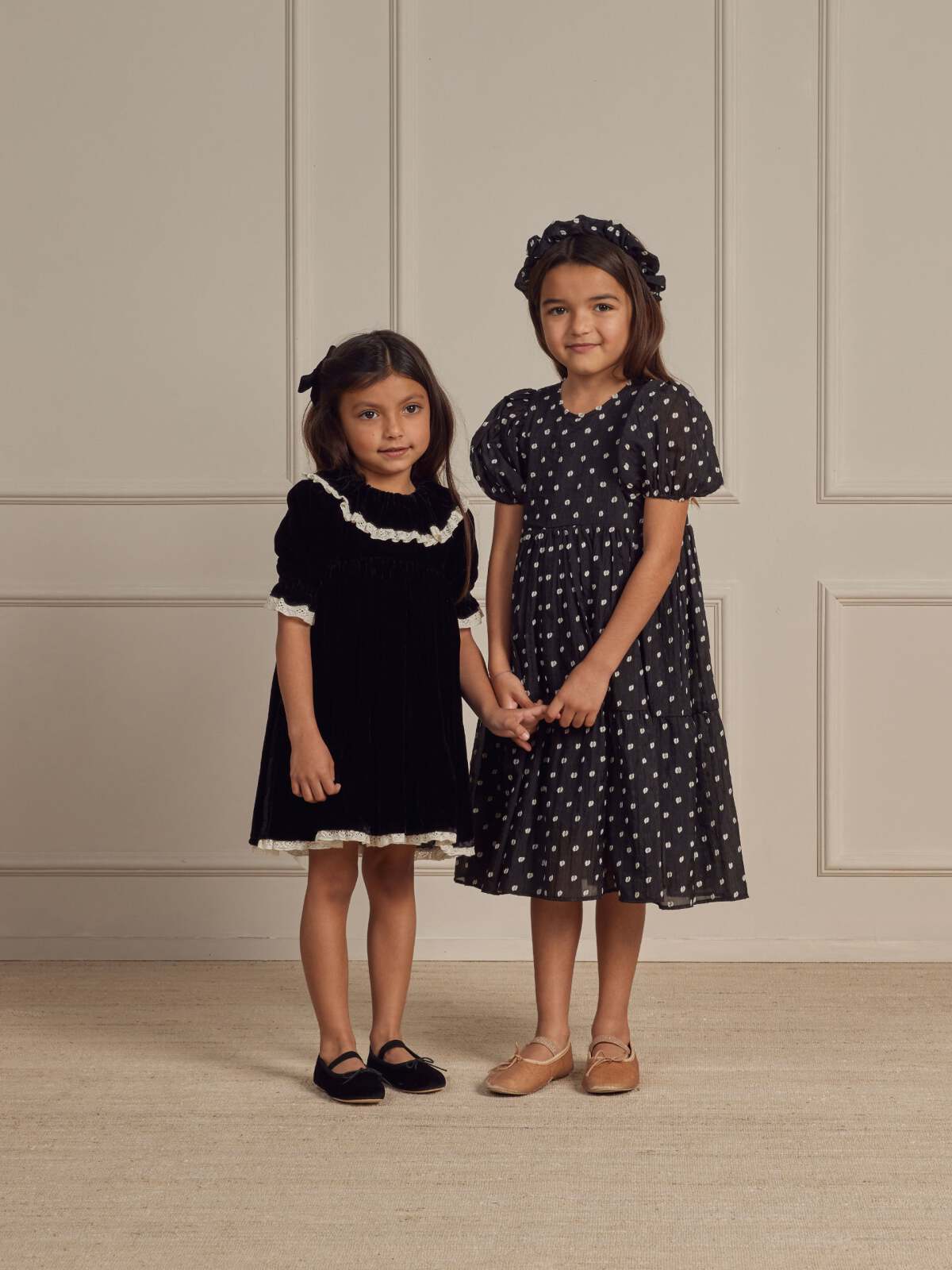 Two young girls in black dresses stand together, holding hands, against a neutral backdrop.