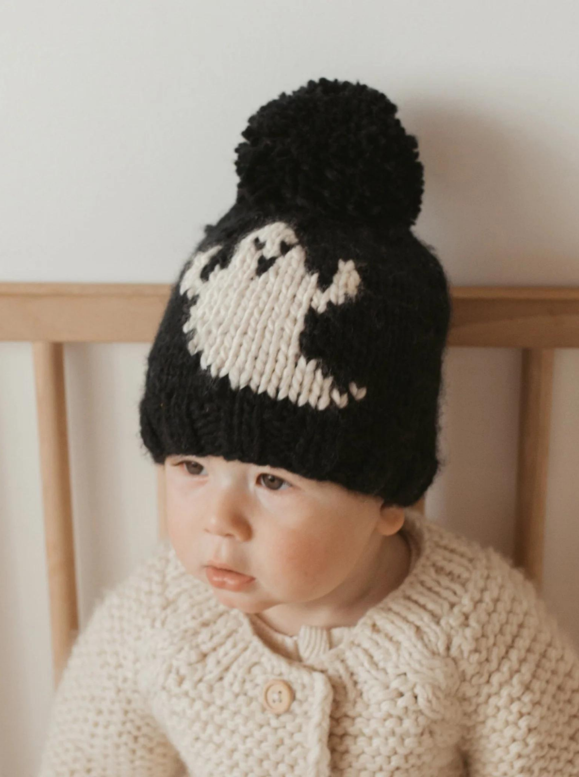 Baby wearing a knitted black hat with a ghost design and a pom-pom, sitting in a cozy sweater.