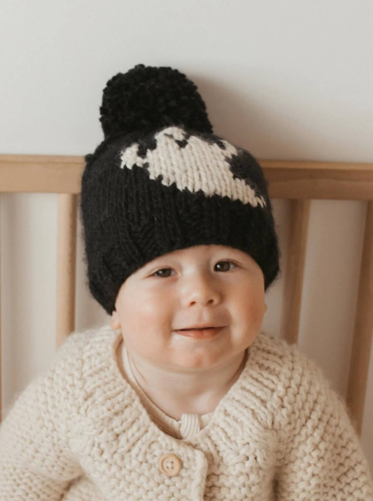 Smiling baby wearing a black knitted hat with a pom-pom and a cream cardigan, seated in a wooden crib.