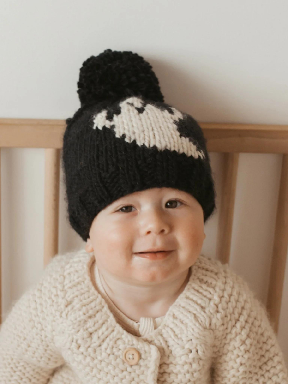 Smiling baby wearing a black knitted hat with a pom-pom and a cream cardigan, seated in a wooden crib.