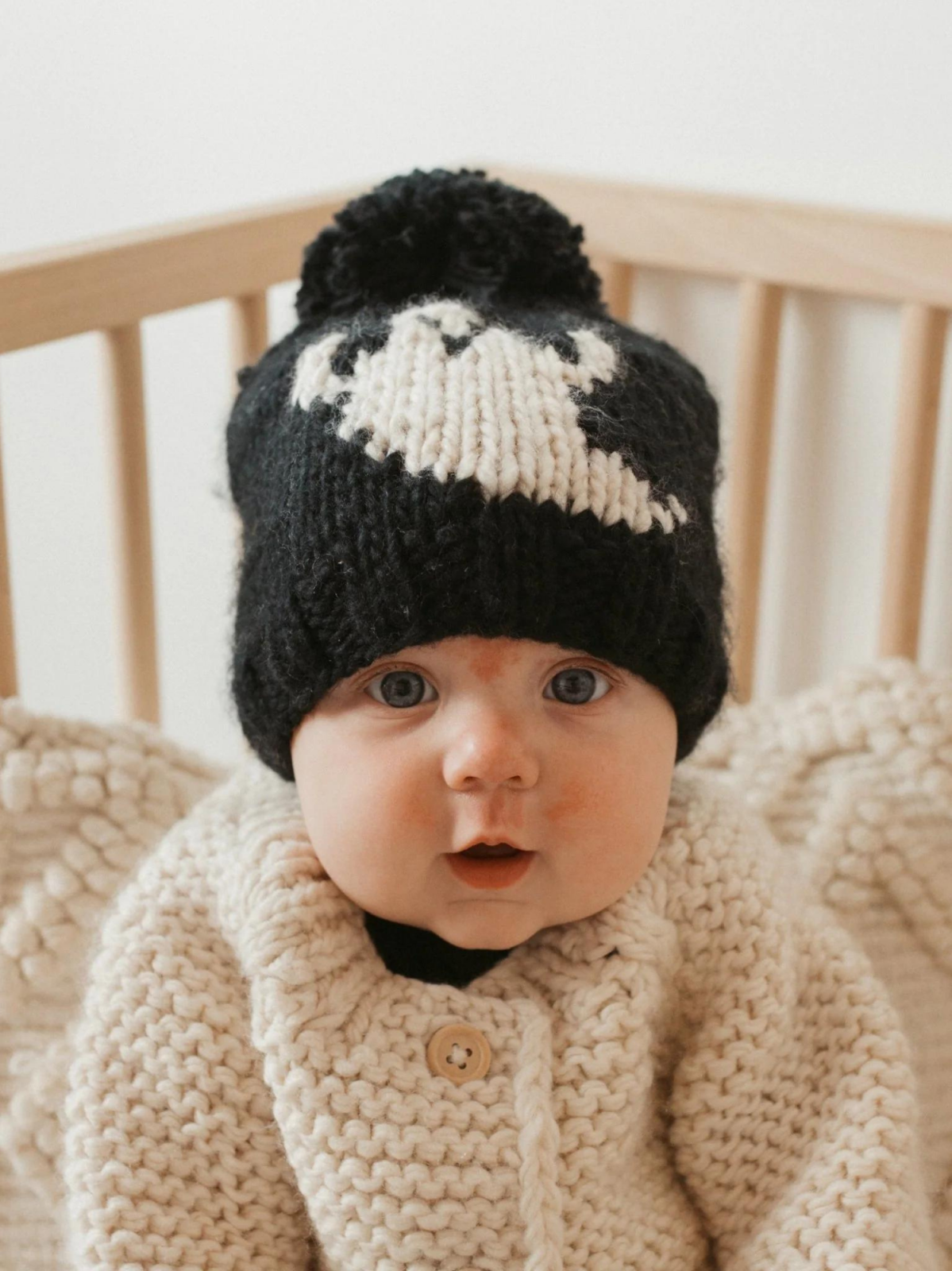 Baby wearing a black and white knit hat, smiling while seated in a wooden crib, dressed in a cozy cream-colored sweater.