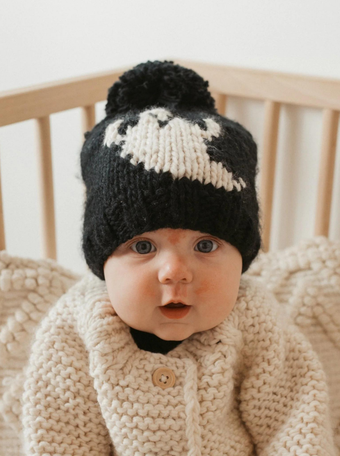 Baby wearing a black and white knit hat, smiling while seated in a wooden crib, dressed in a cozy cream-colored sweater.