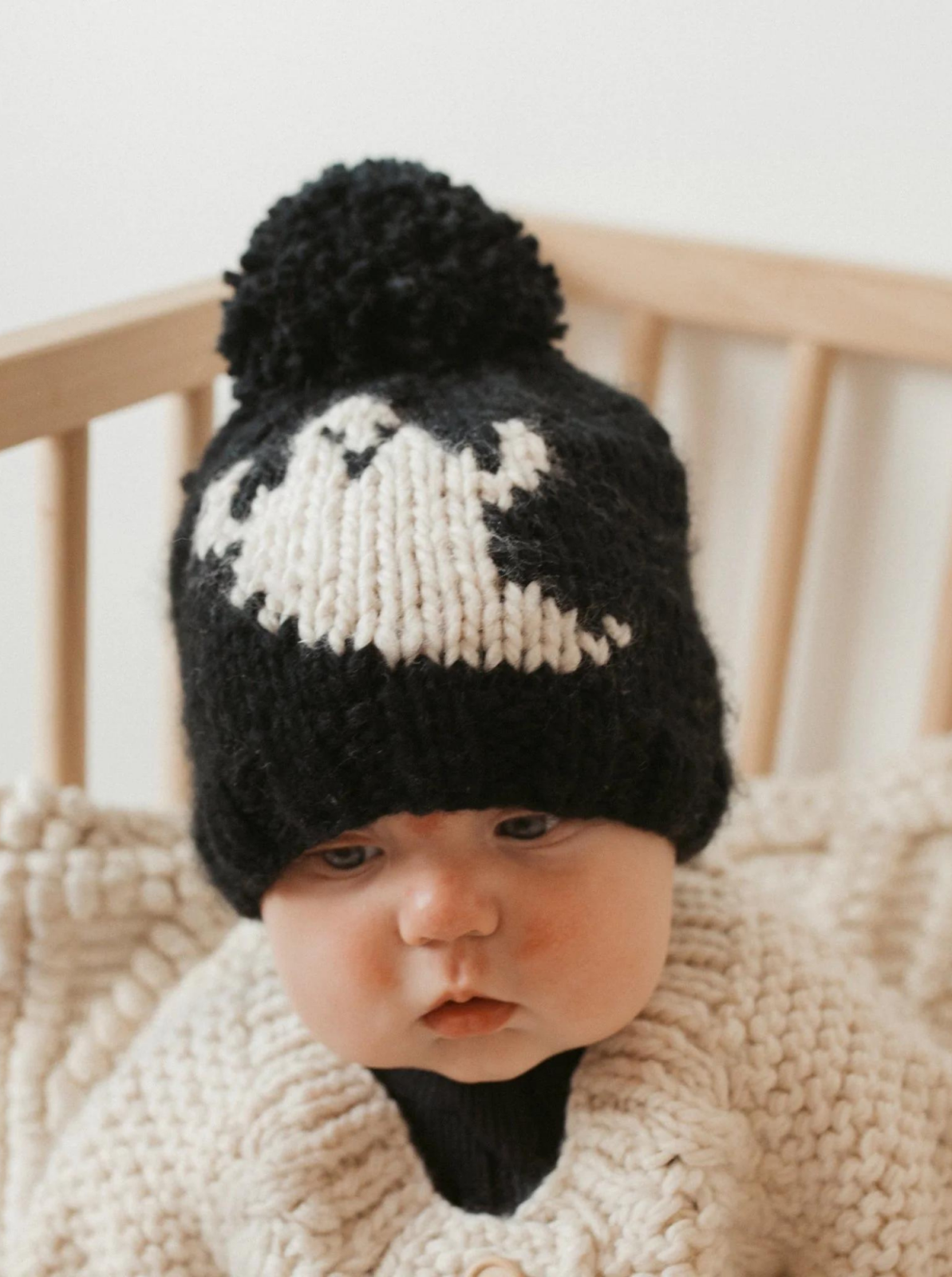 Baby wearing a black and white knitted hat with pompom, sitting in a cozy, knitted blanket in a wooden crib.